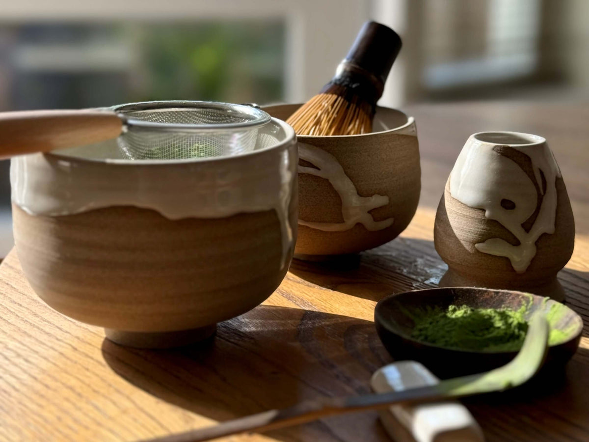 A handmade ceramic matcha set including a chawan with a metal sifter, a bamboo chasen (whisk) in another bowl, and a chashaku (scoop) resting on a small dish with matcha powder — all beautifully lit by soft natural light.