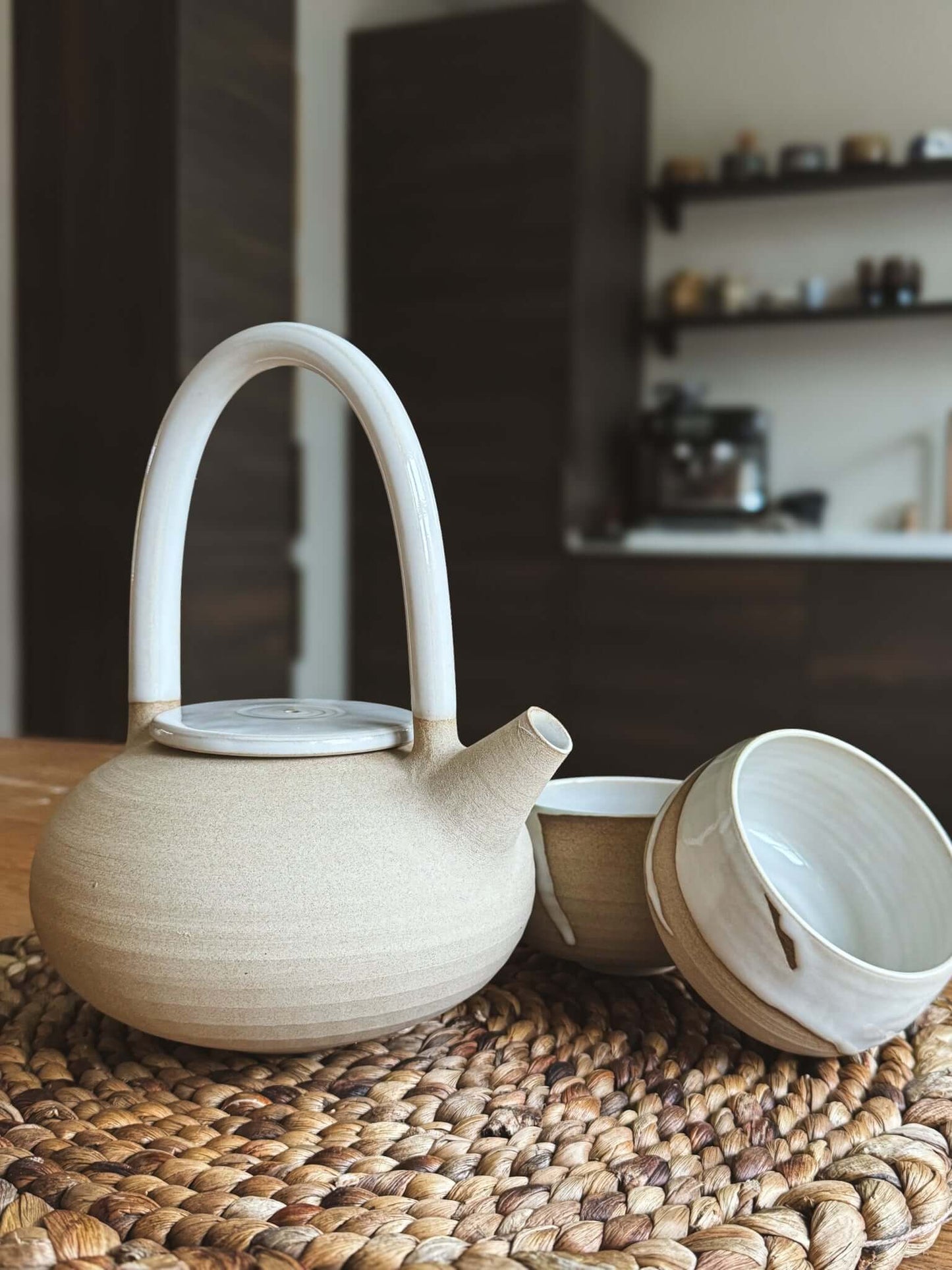 Teapot - Yuki #02 on a woven mat, featuring a tall looped handle, glossy white glazed curves, and unglazed warm clay lower half.