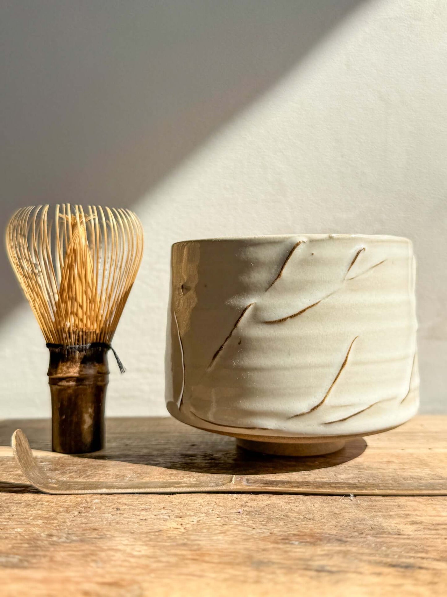 Ivory matcha bowl with delicate slip lines, part of the Yuki Series, beside a bamboo whisk and scoop on a wooden table.