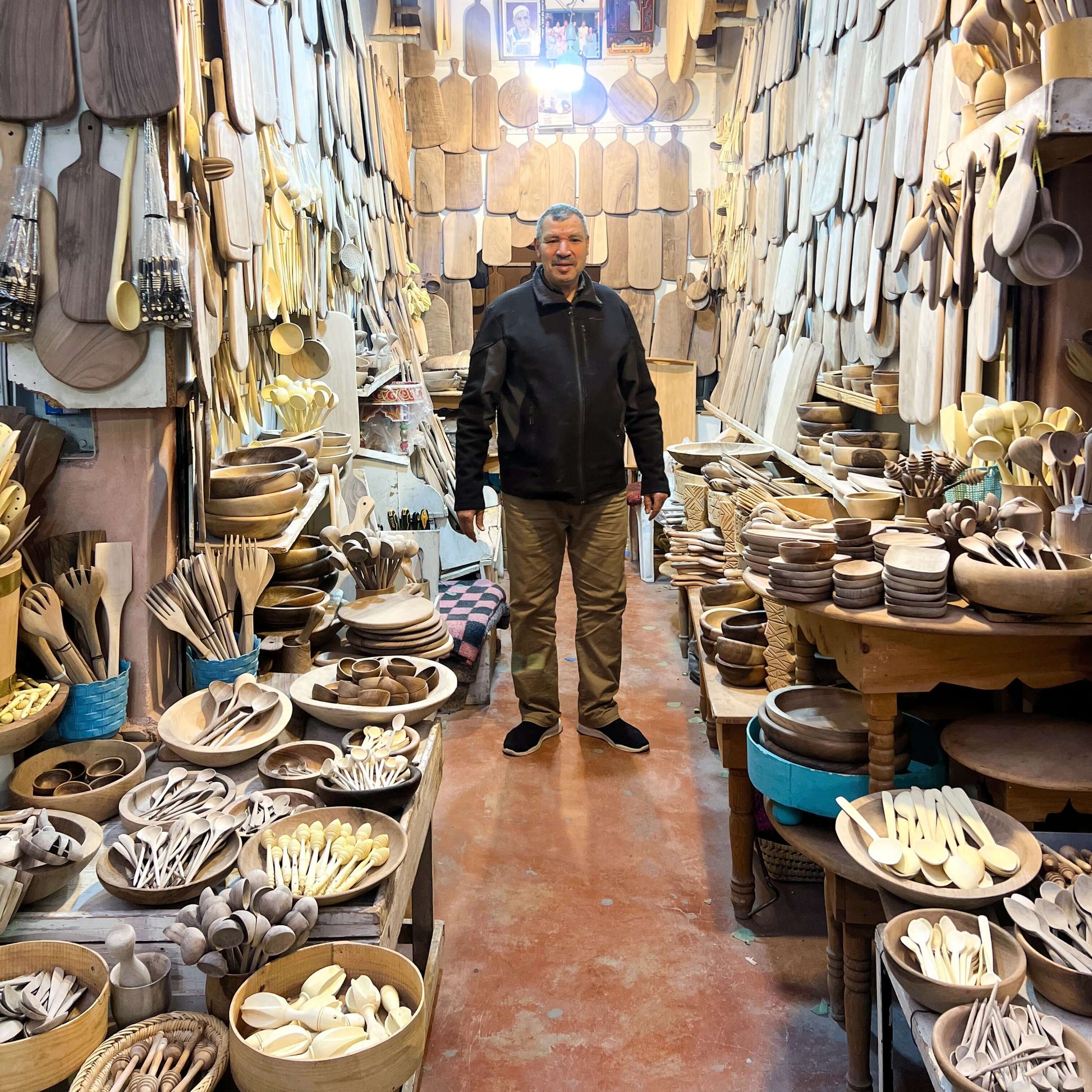 Artisan in shop displaying hand-carved walnut tea spoons and handmade ceramic items in Marrakech Medina.