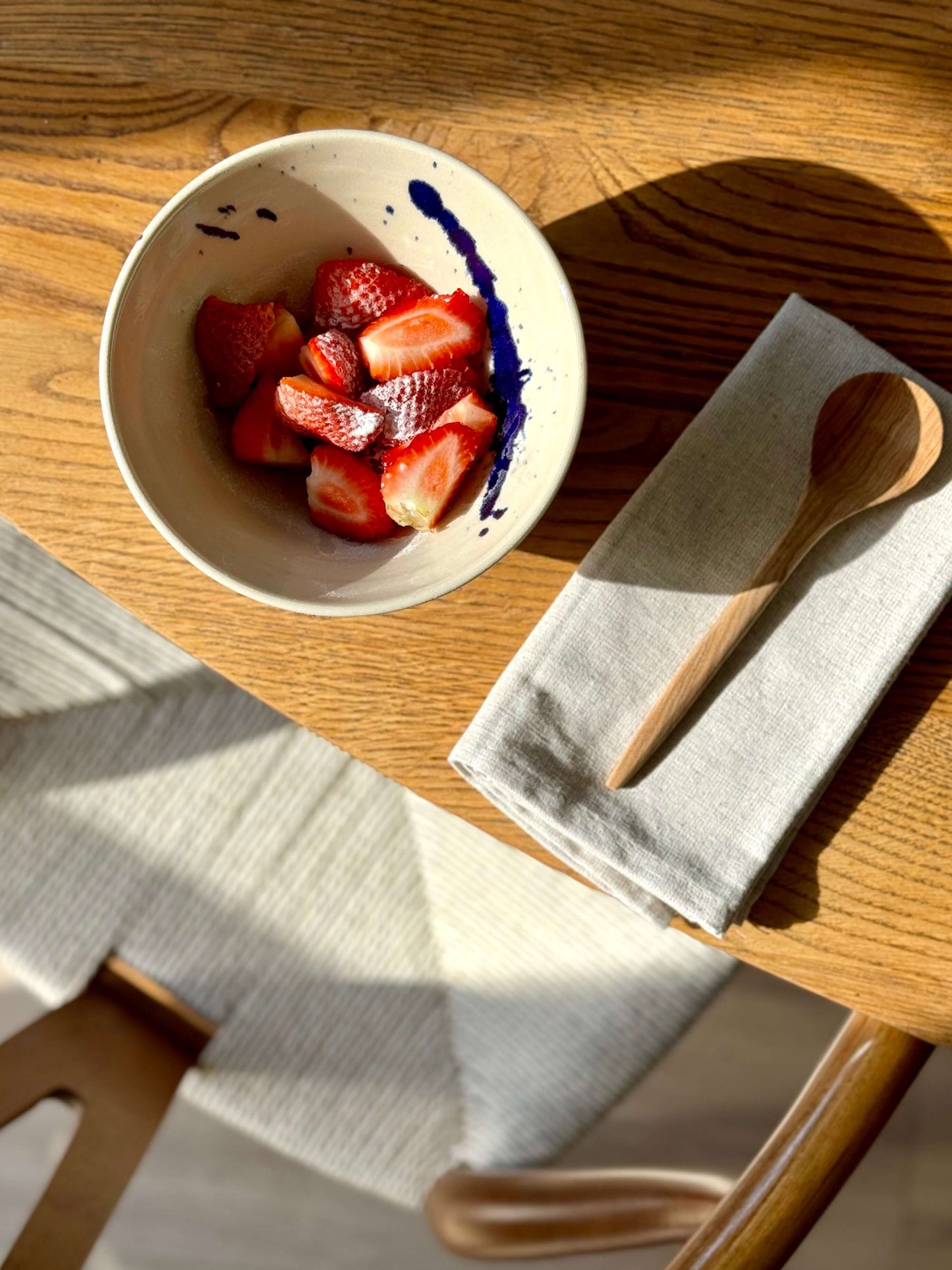Ceramic breakfast bowl with strawberries and cobalt blue accents on a wooden table next to a linen napkin and wooden spoon.