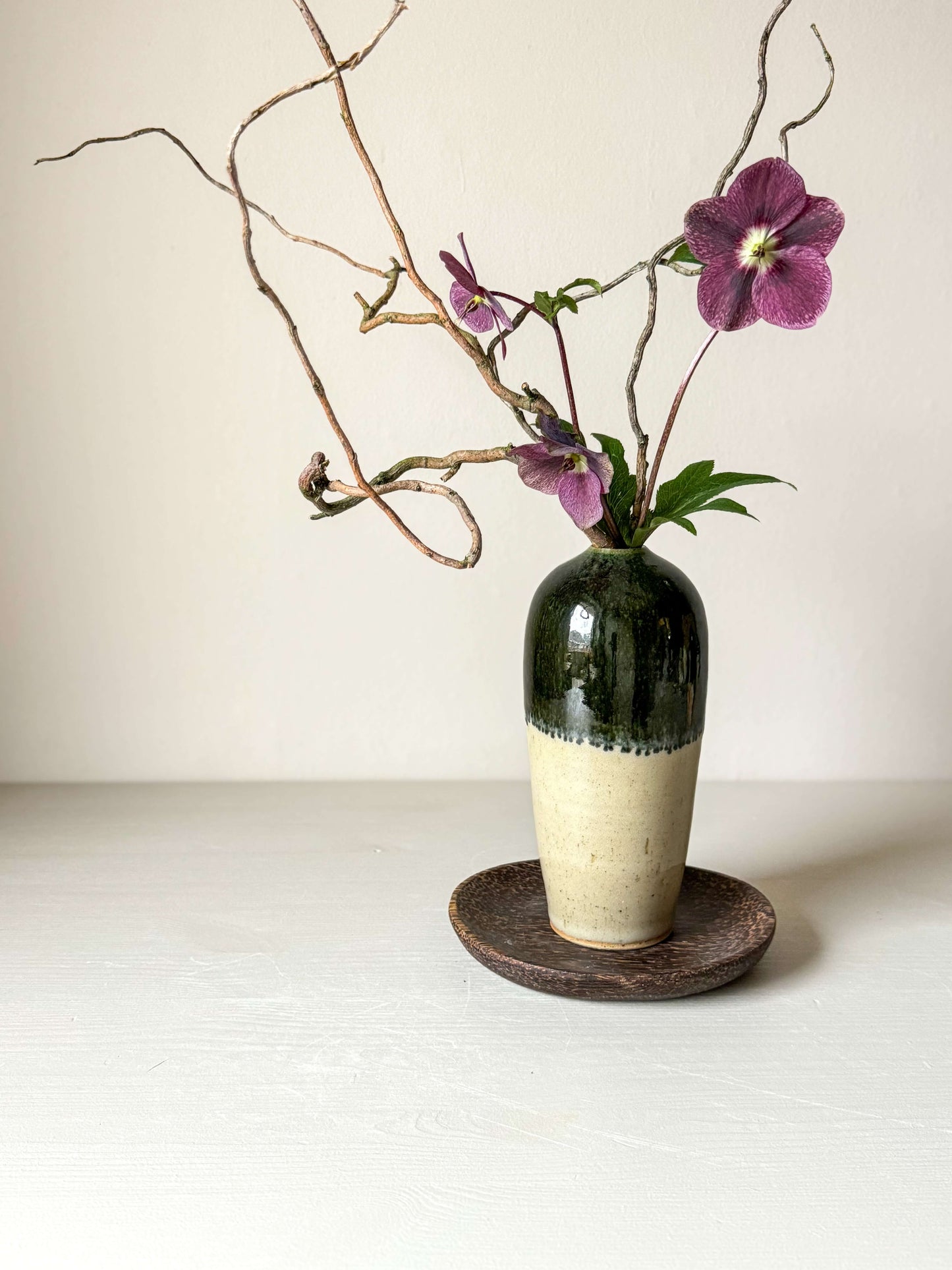 Handcrafted ceramic vase with a glossy finish and green rim, adorned with branches and purple flowers, sitting on a wooden coaster.