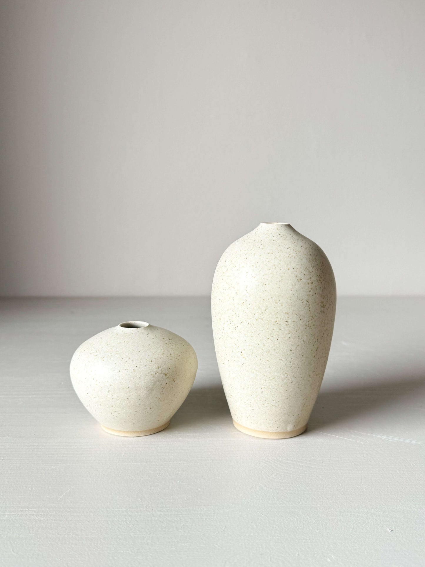 Two ceramic vases with speckled matte cream glaze, featuring a softly tapered shape and narrow openings, displayed on a neutral background.