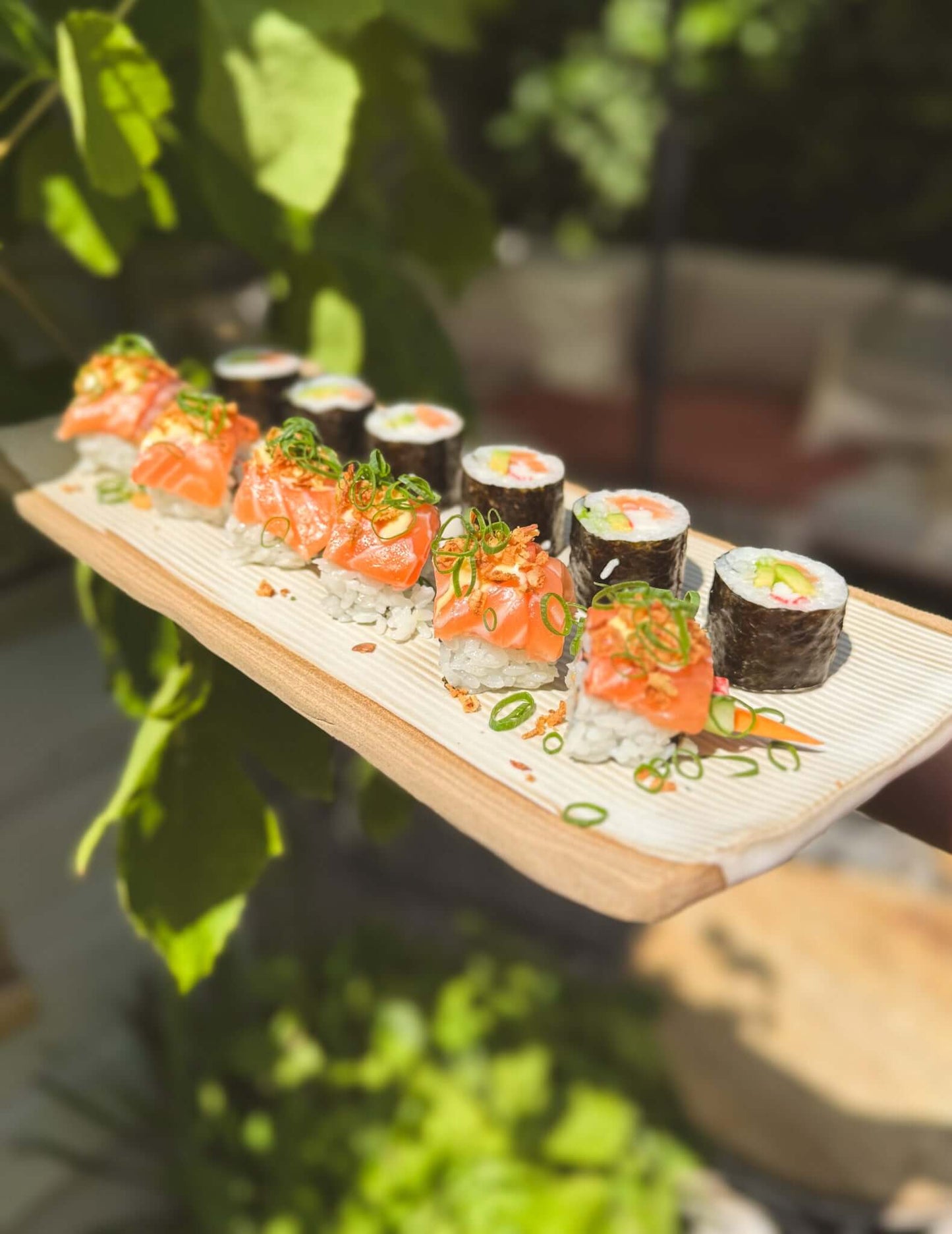 Assorted sushi rolls with vibrant toppings served outdoors on a wooden platter, surrounded by lush greenery.