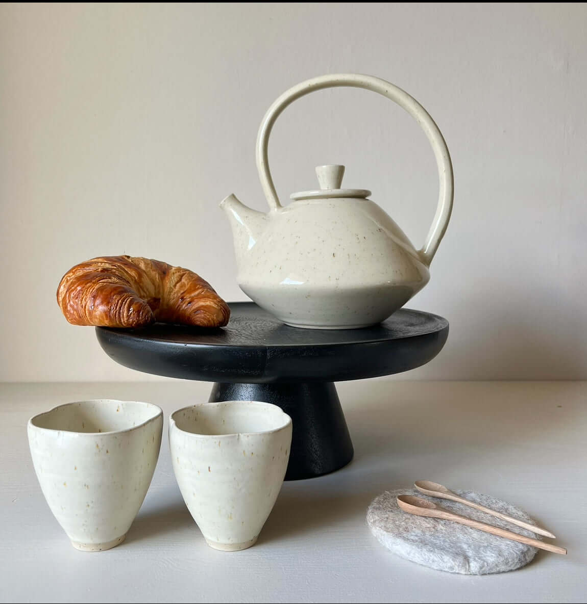 Hand-Carved Walnut Tea Spoons on a marble tray with a handmade ceramic teapot, cups, and a croissant on a black pedestal.