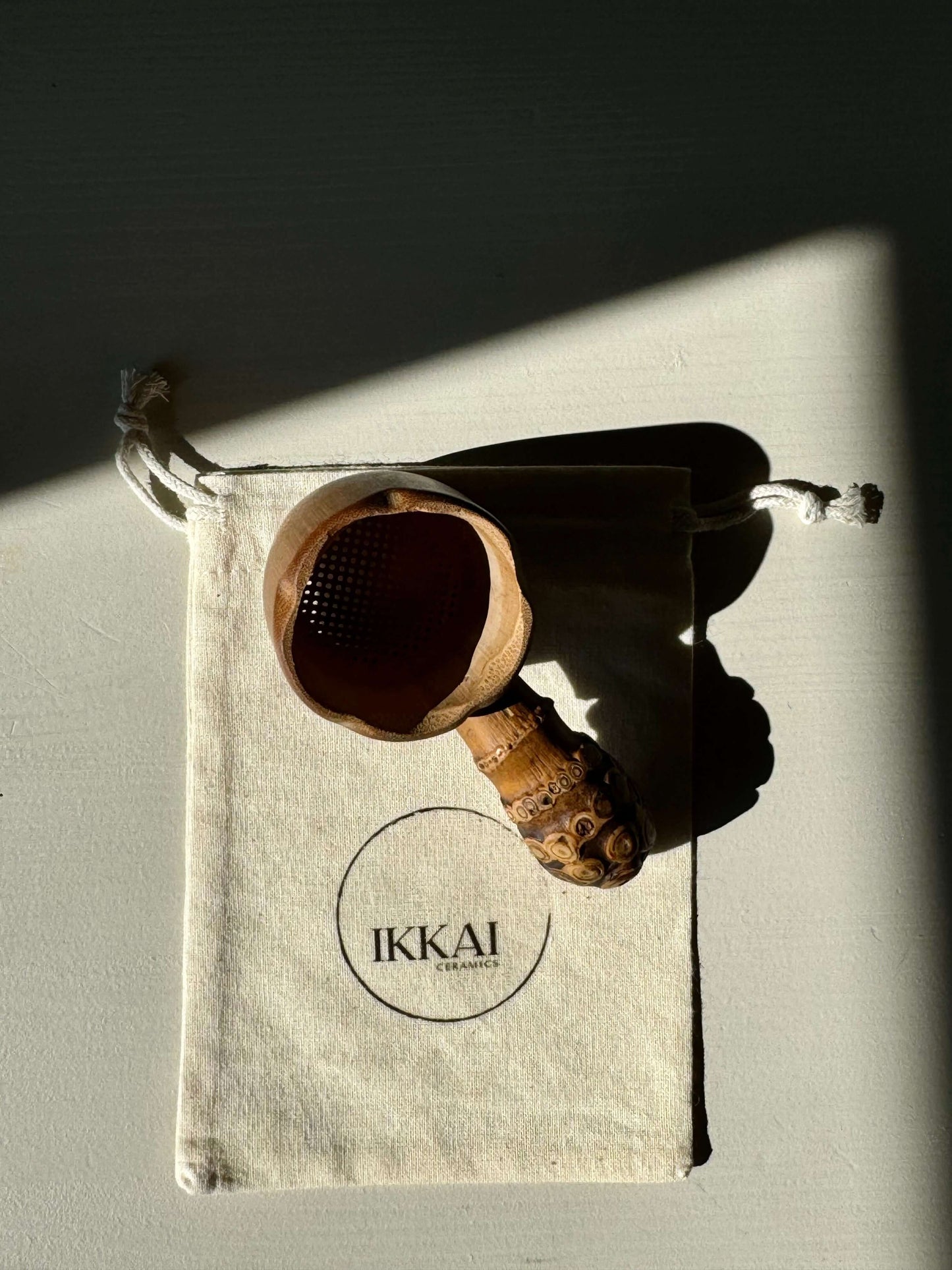 Handmade bamboo tea strainer with rustic root handle on a branded IKKAI drawstring bag.