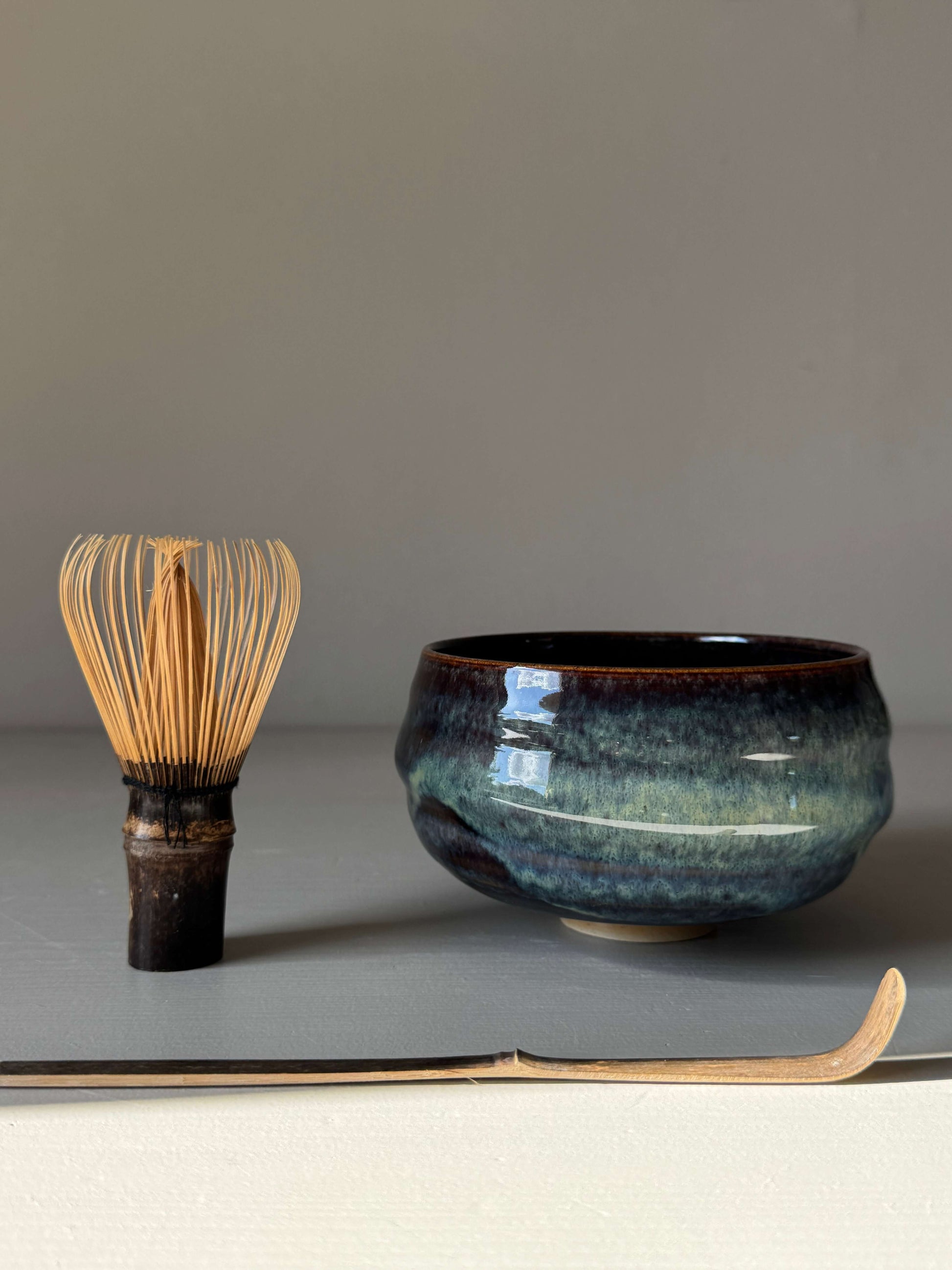 Matcha Bowl - Glossy Blue #03, handmade ceramic with flowing curves next to a bamboo whisk and scoop, earthy tones and unpredictable blues