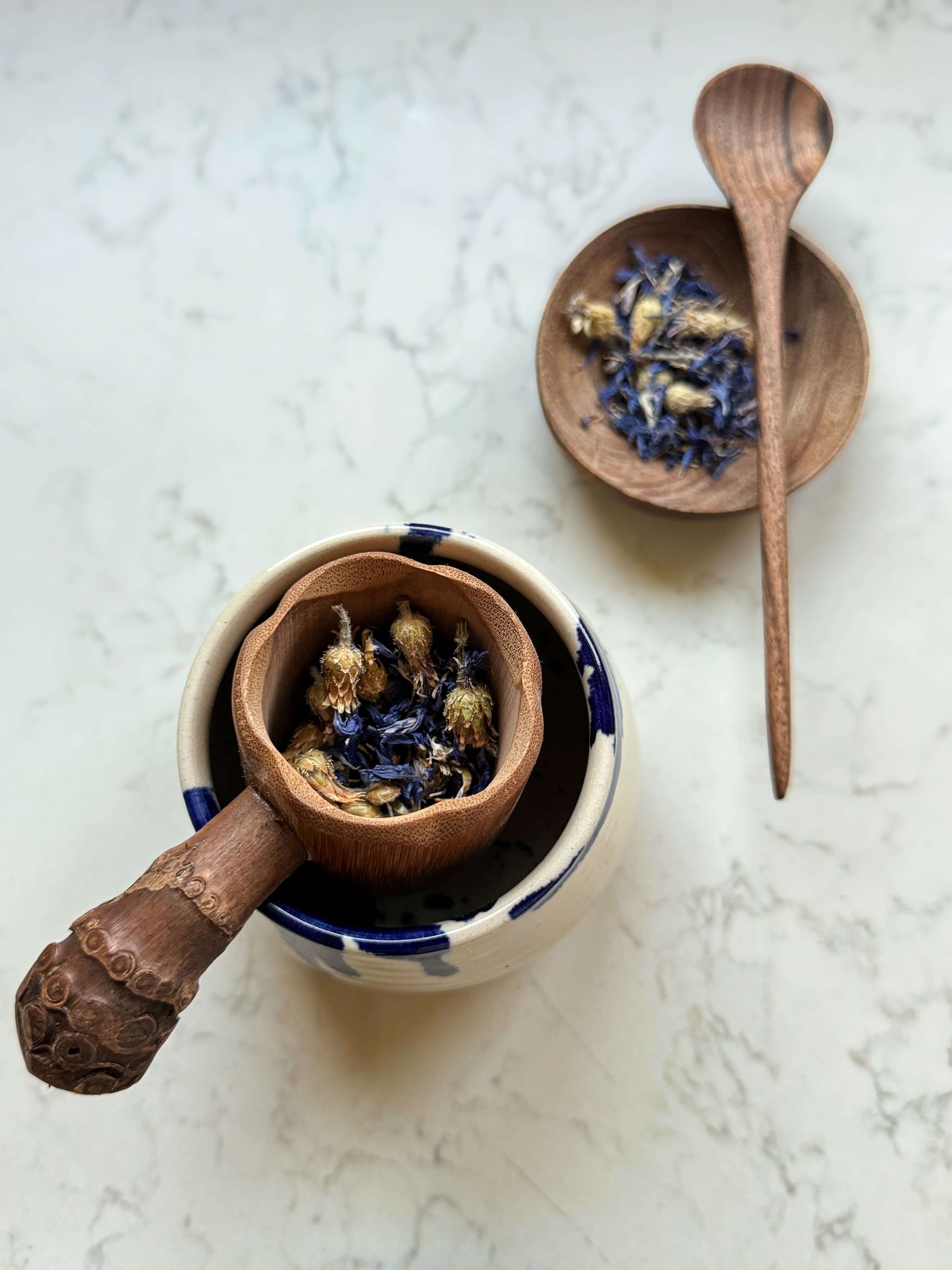 Handmade bamboo tea strainer with root handle placed on a mug, featuring dried flowers, next to a wooden spoon and bowl.
