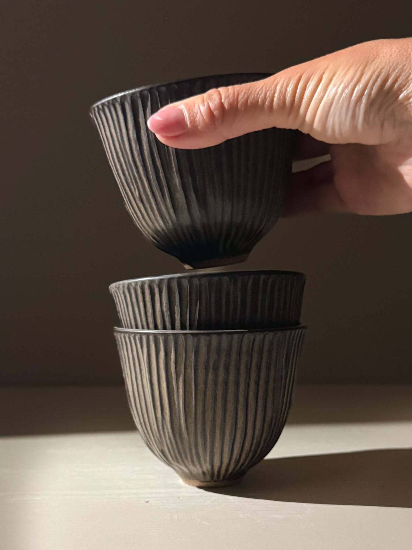 Hand holding a stack of three handmade ceramics, Carved Yunomi Cup - Black Truffle, showcasing pottery texture and vertical lines.