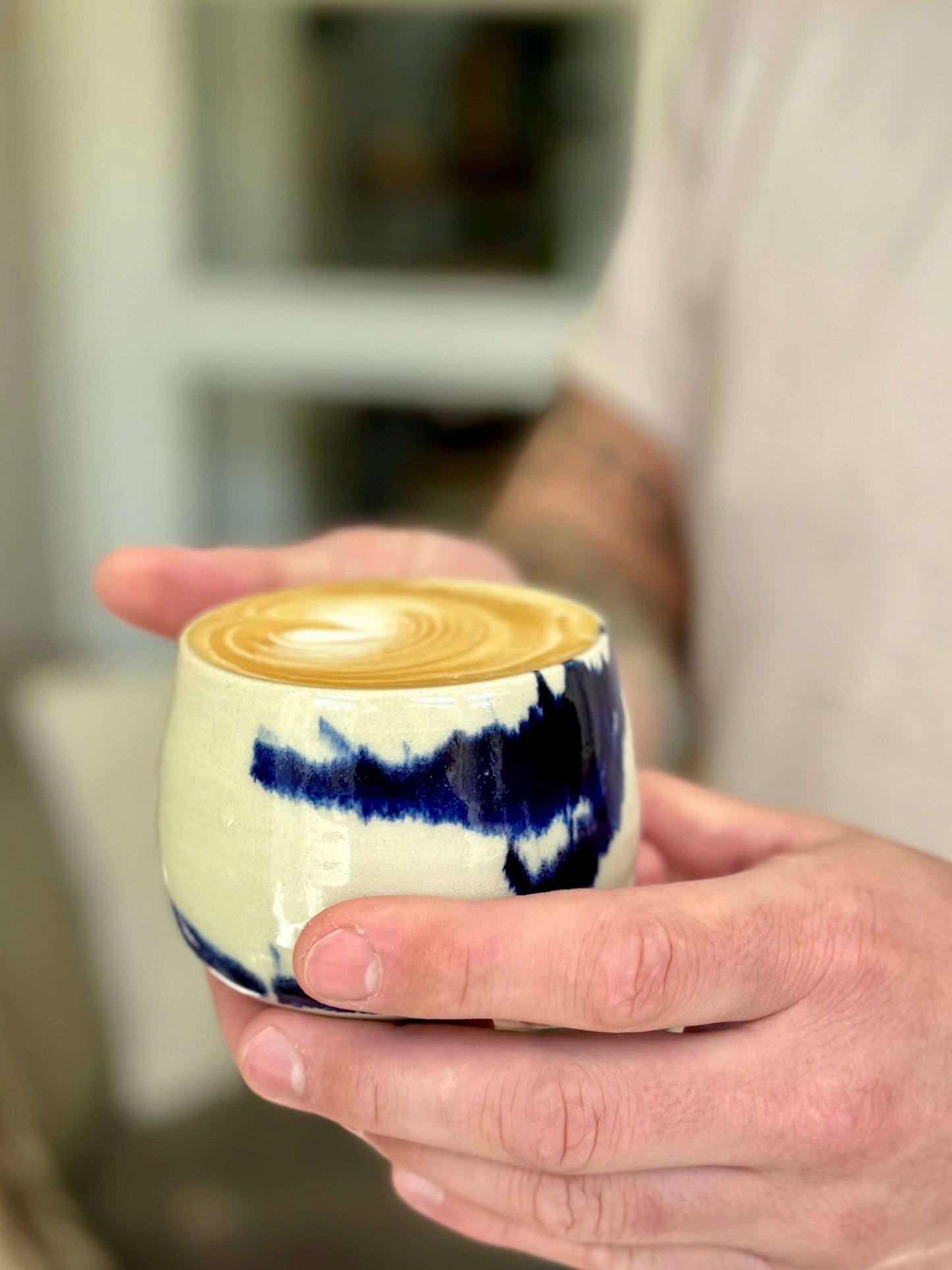 Hand holding a ceramic coffee cup with blue splashes from the Gosu Series, filled with latte art.