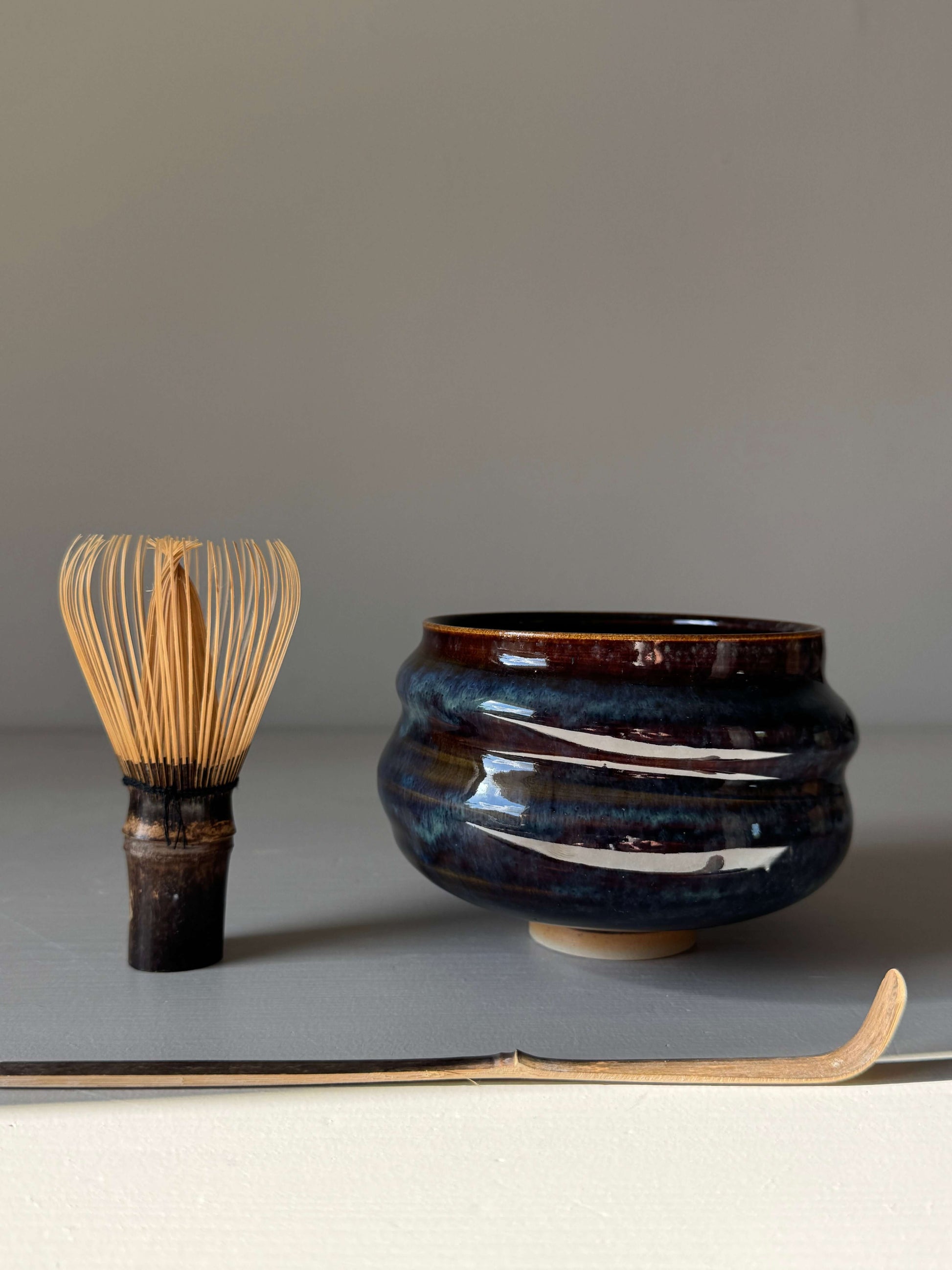 Matcha Bowl - Glossy Blue #04, handmade ceramic bowl with unique earthy brown and flowing blue hues, next to a matcha whisk.