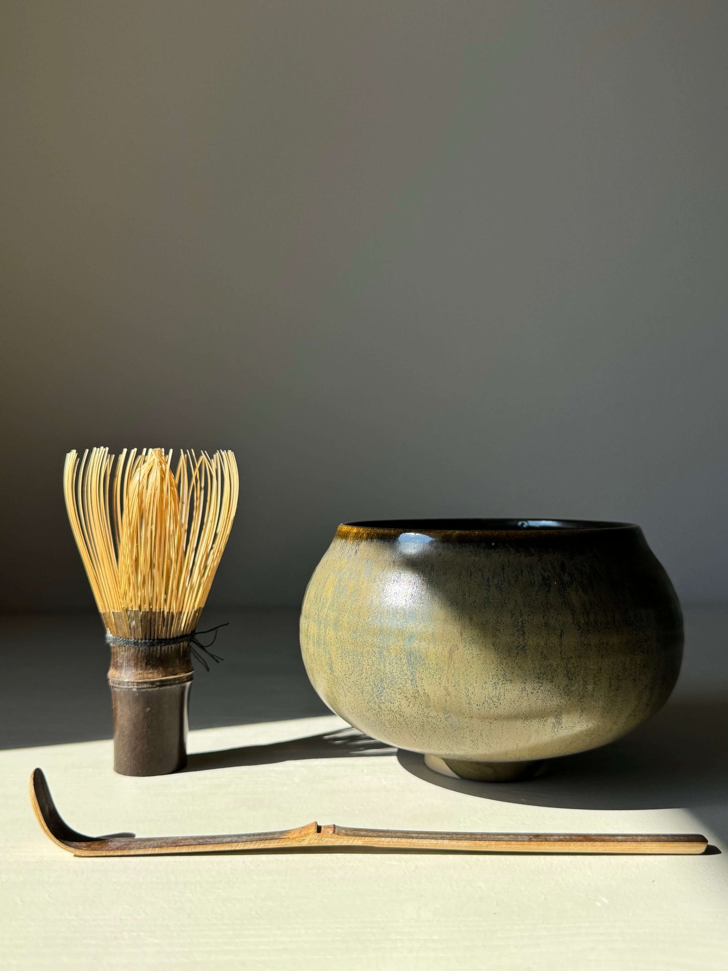 Sabi Black Matcha Bowl with bamboo whisk and scoop in soft light, featuring golden ochre and subtle blue-green tones.