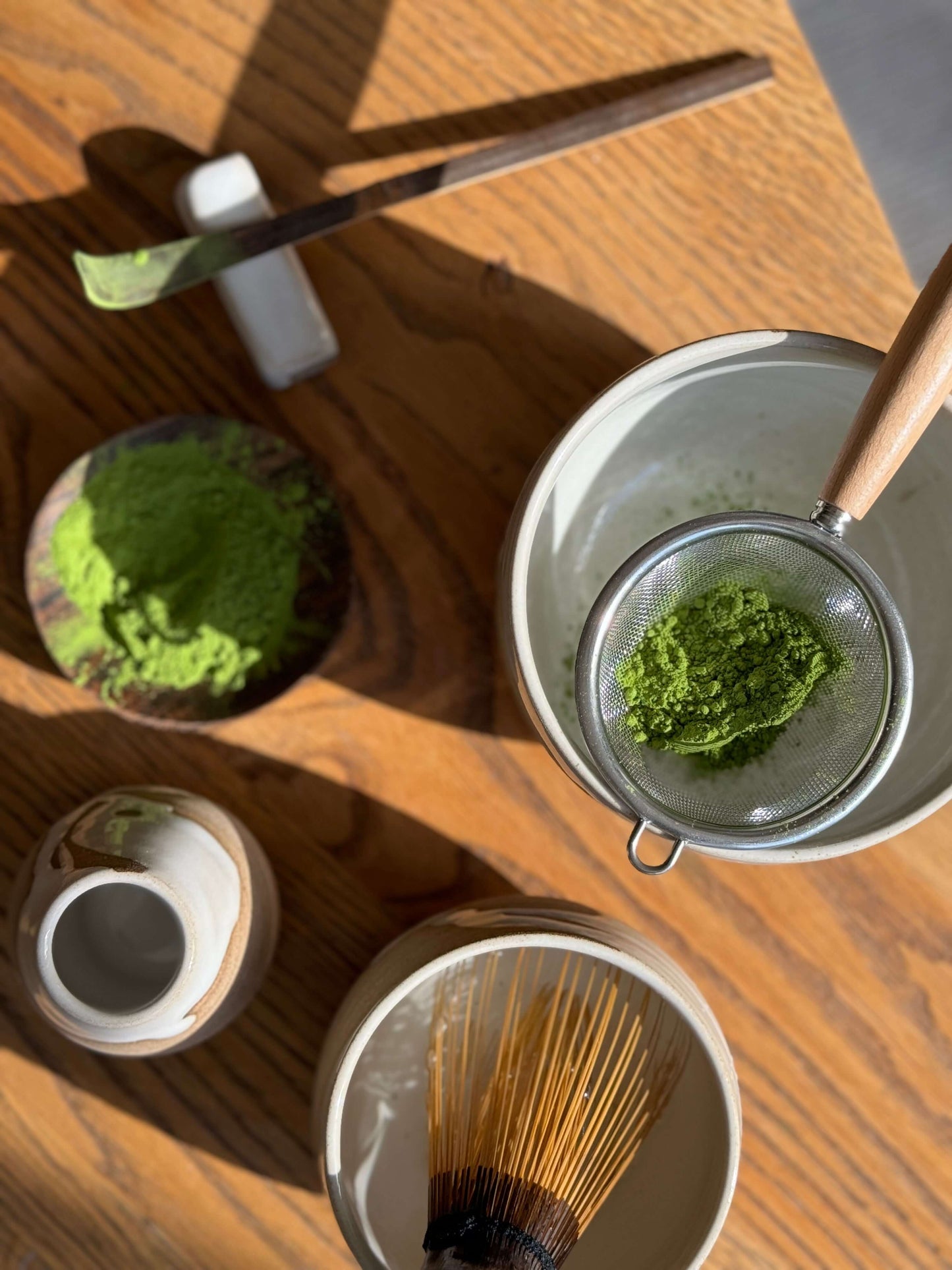 Top view of a Matcha tea set with a Yuki Series Matcha bowl, bamboo whisk, green matcha powder and sifter on a wooden table.