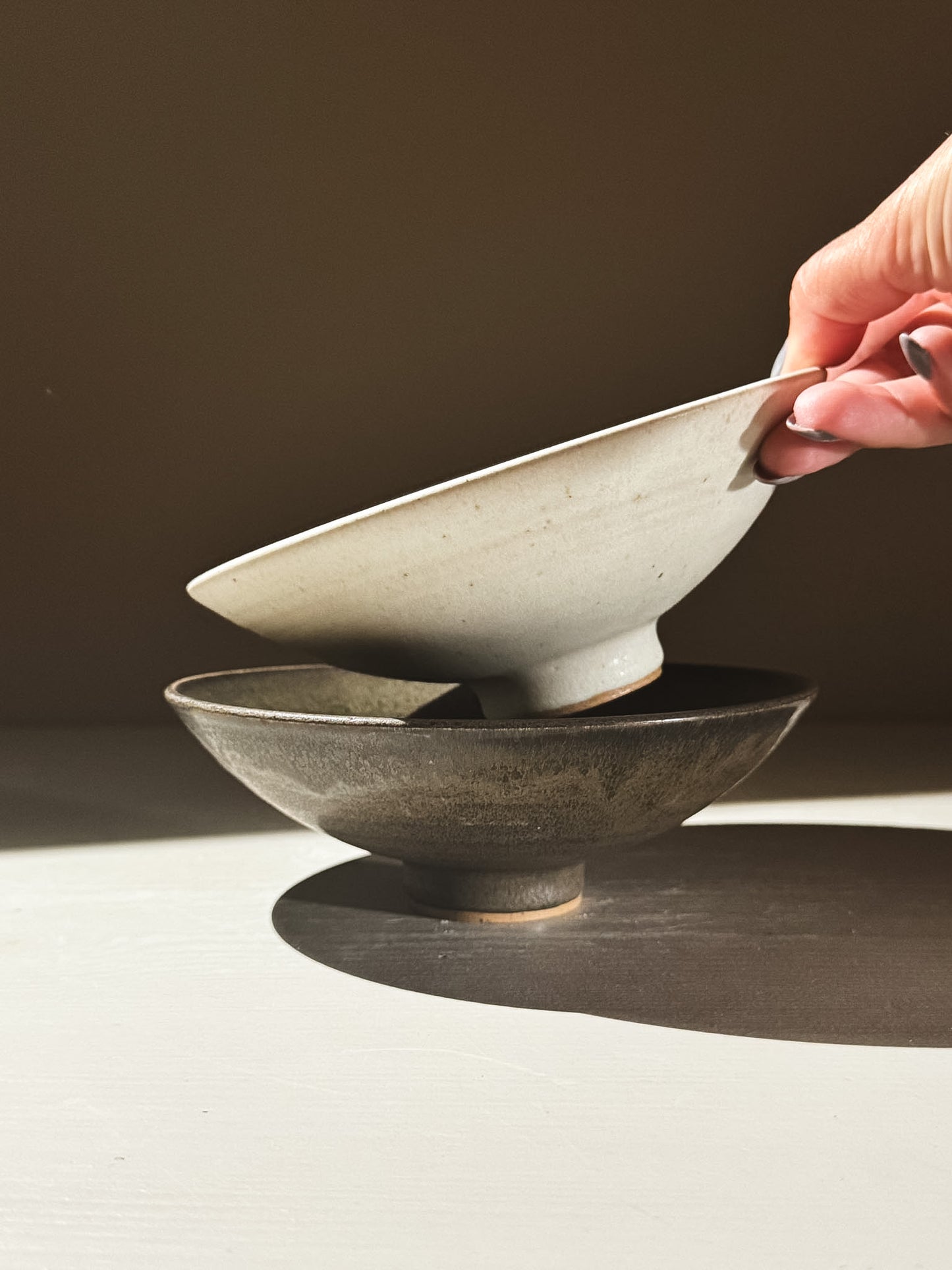 Small Footed Serving Bowl – Eggshell being held above a dark bowl, showcasing its elegant shape and matte finish.