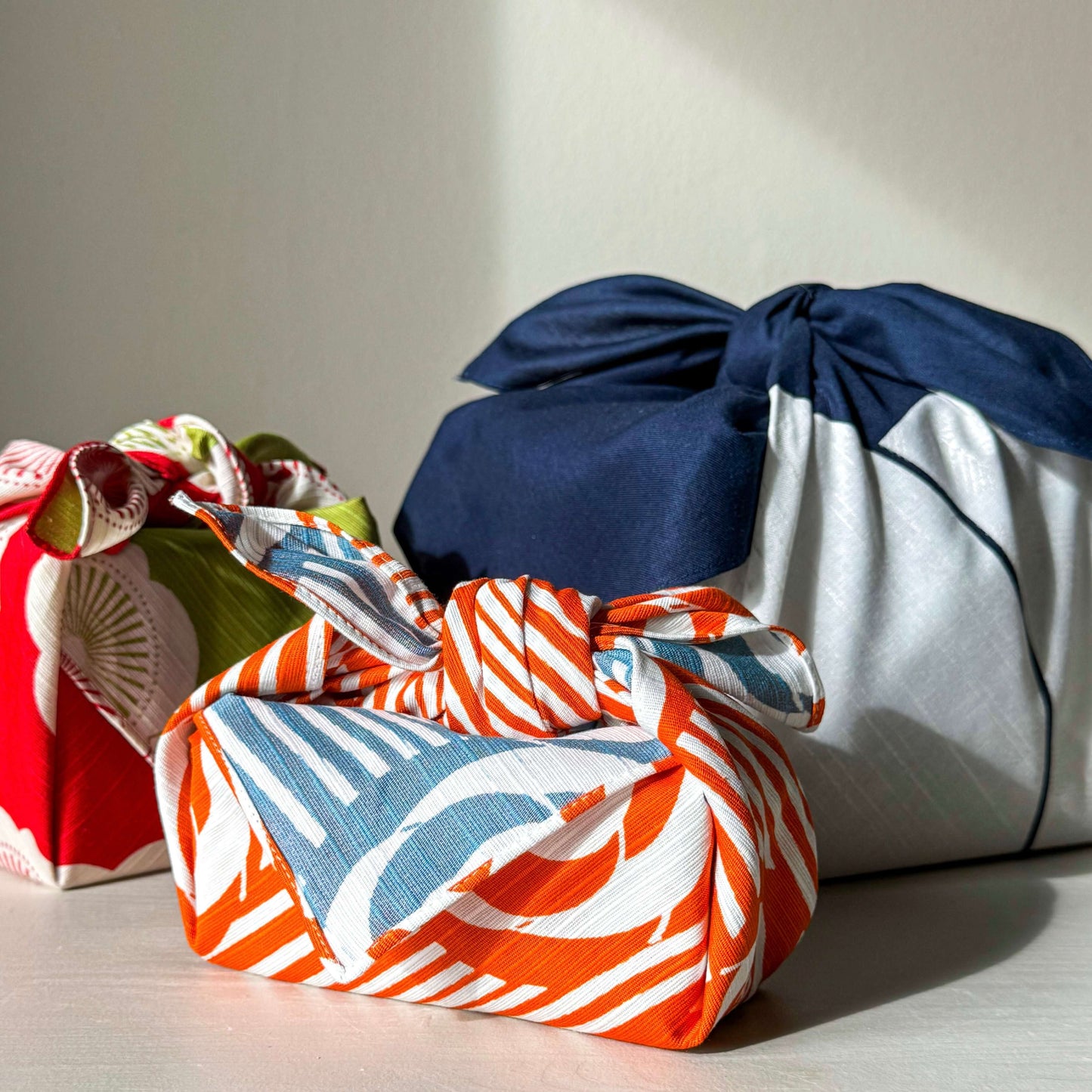 Small Furoshiki in Camellia Orange & Blue with a floral pattern, elegantly wrapped next to other Furoshiki designs, in soft lighting.