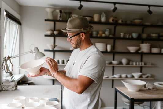 Abel pepping in home pottery studio inspecting a handmade ceramic bowl, with shelves of bisqueware and pottery tools in the background.