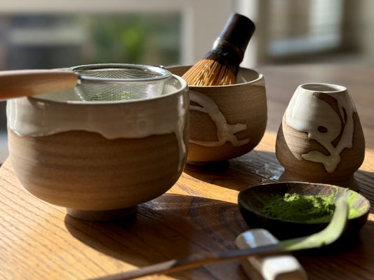 A handmade ceramic matcha set including a chawan with a metal sifter, a bamboo chasen (whisk) in another bowl, and a chashaku (scoop) resting on a small dish with matcha powder — all beautifully lit by soft natural light.