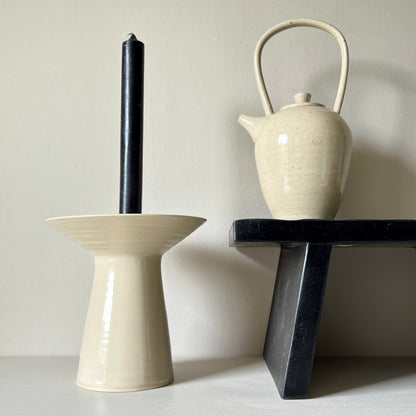 A large cream ceramic teapot standing on a black bench next to a cream ceramic candle holder holding a black candle