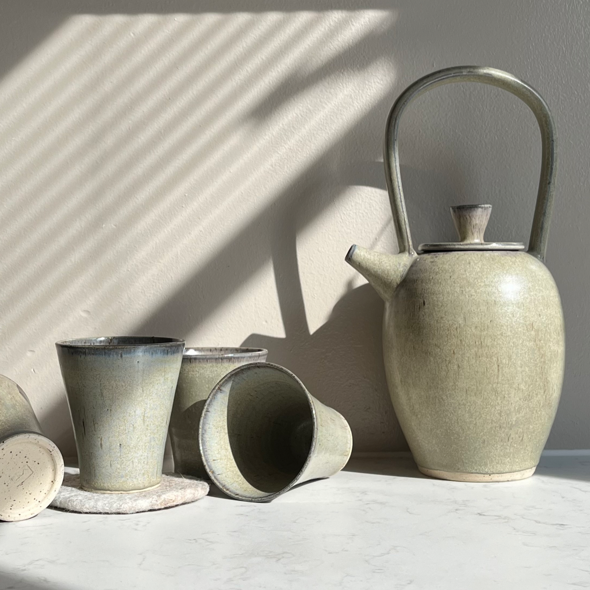 Blue ceramic teapot on a marble table, accompanied by three blue ceramic tumblers, set against a neutral background