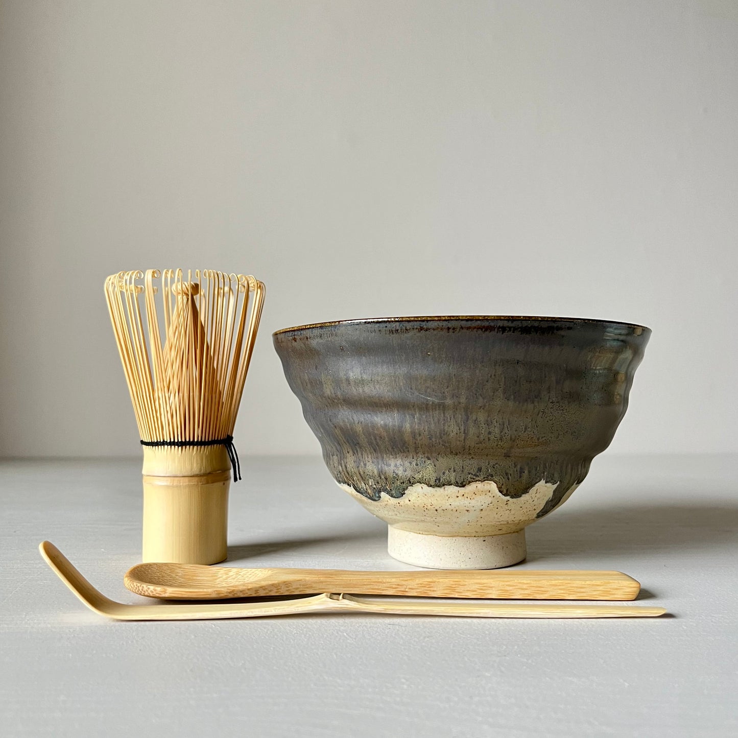 A black and cream handmade ceramic matcha bowl standing next to a matcha whisk with another bamboo matcha spoon in the foreground