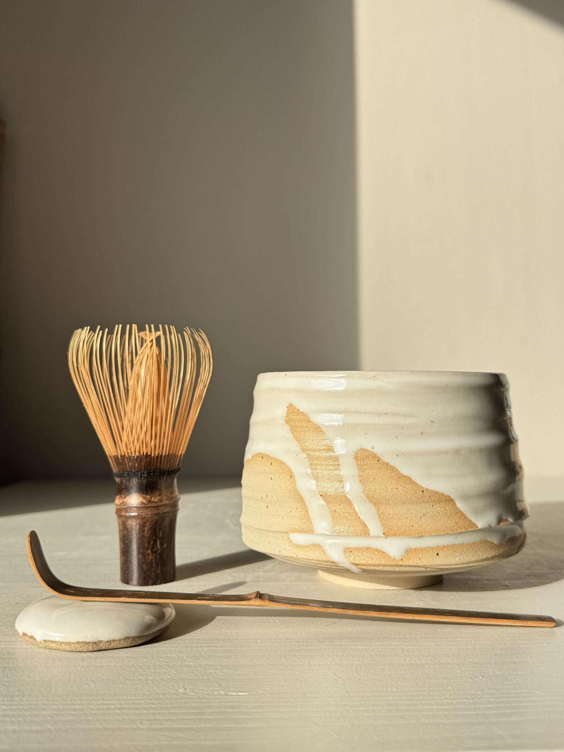 Matcha Bowl – Half Glossy White Glaze #01, handmade ceramic with speckled stoneware clay, beside a whisk and tea scoop.