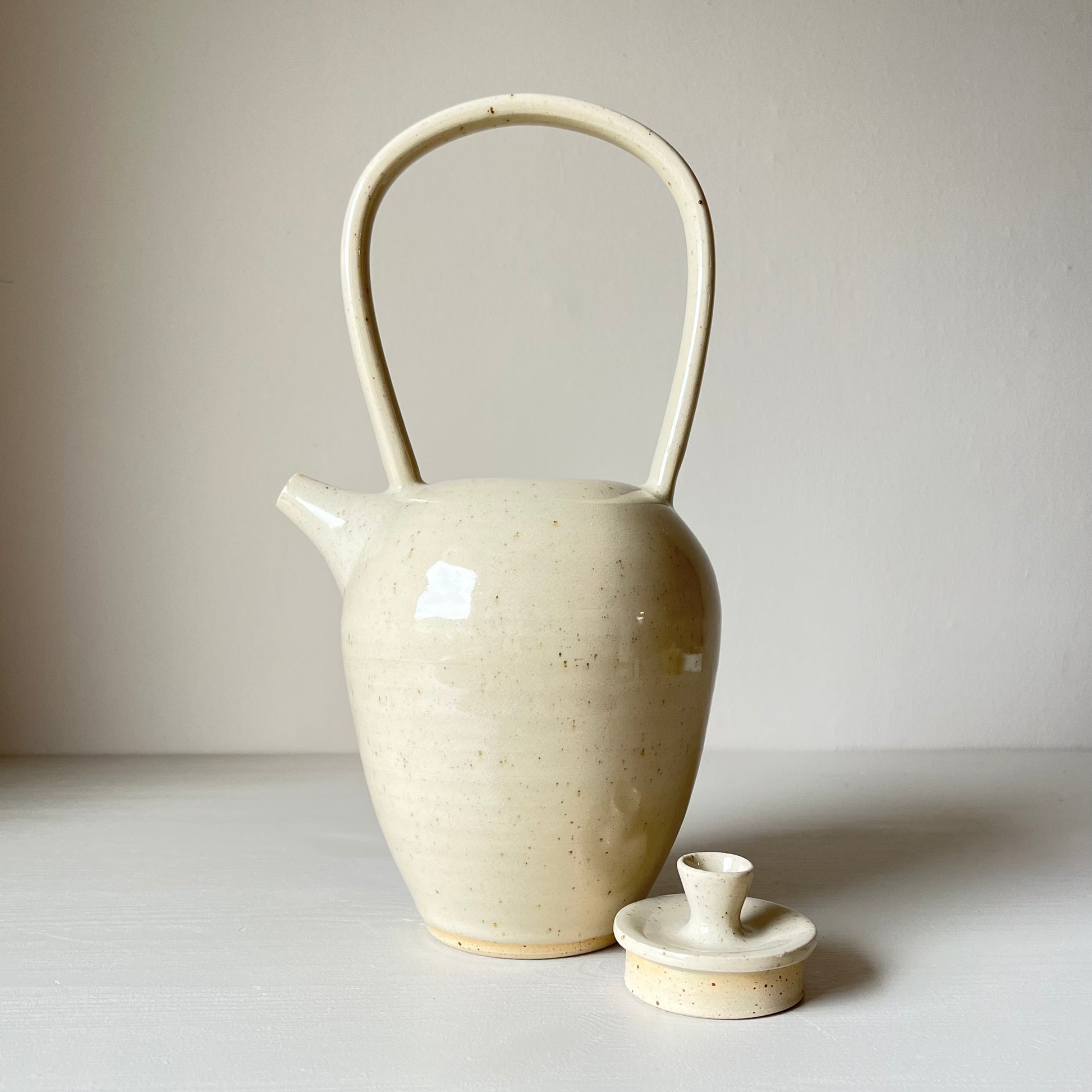 a tall ceramic teapot and lid with a cream coloured glaze on a neutral background