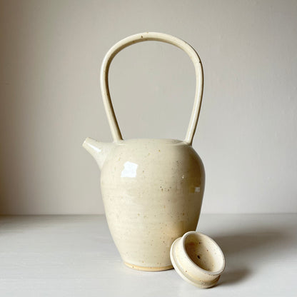 a tall ceramic teapot and lid with a cream coloured glaze on a neutral background