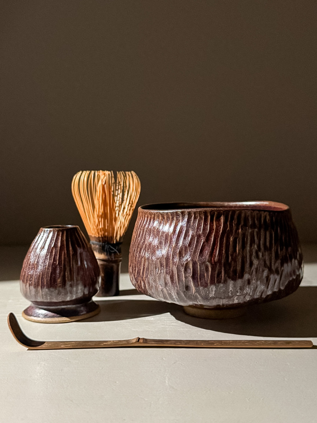 A set of four handmade ceramic matcha tea tools, including a large textured bowl, a small vase, a whisk, and a bamboo scoop, arranged on a neutral surface against a dark background.