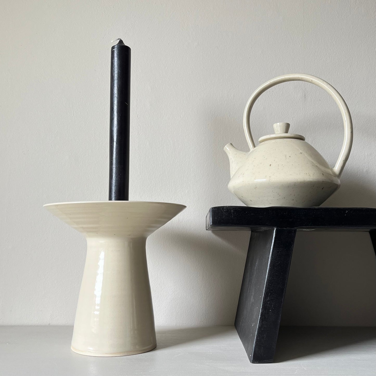 A large cream ceramic teapot on a black bench, next to another large cream ceramic candlestick holder and a black candlestick.