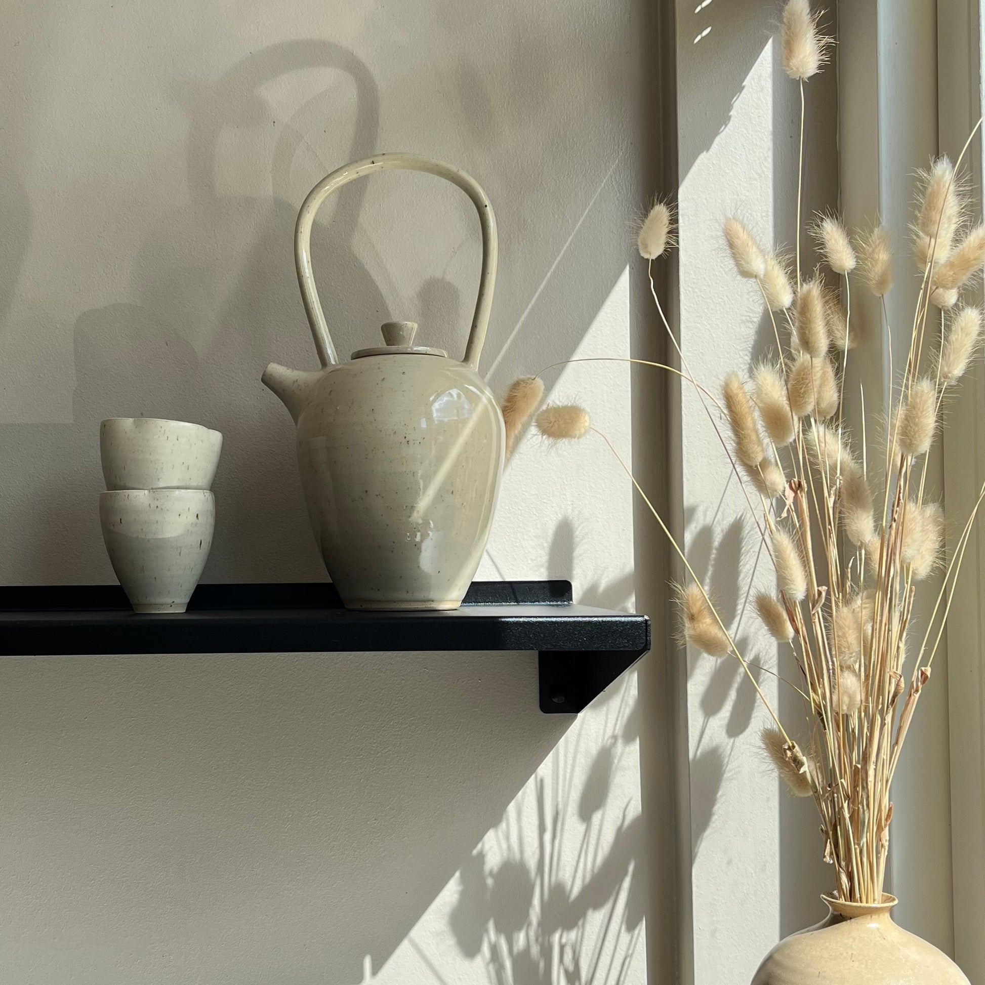 A large ceramic teapot on a black shelf, next to two small cups and a peach vase with dried flowers.