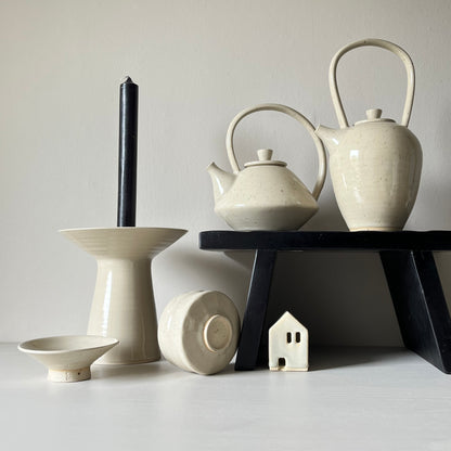  Two cream ceramic teapots standing on a small black bench with a small ceramic house underneath, next to a large white ceramic candleholder, a white matcha bowl, and a white ceramic soap dish.