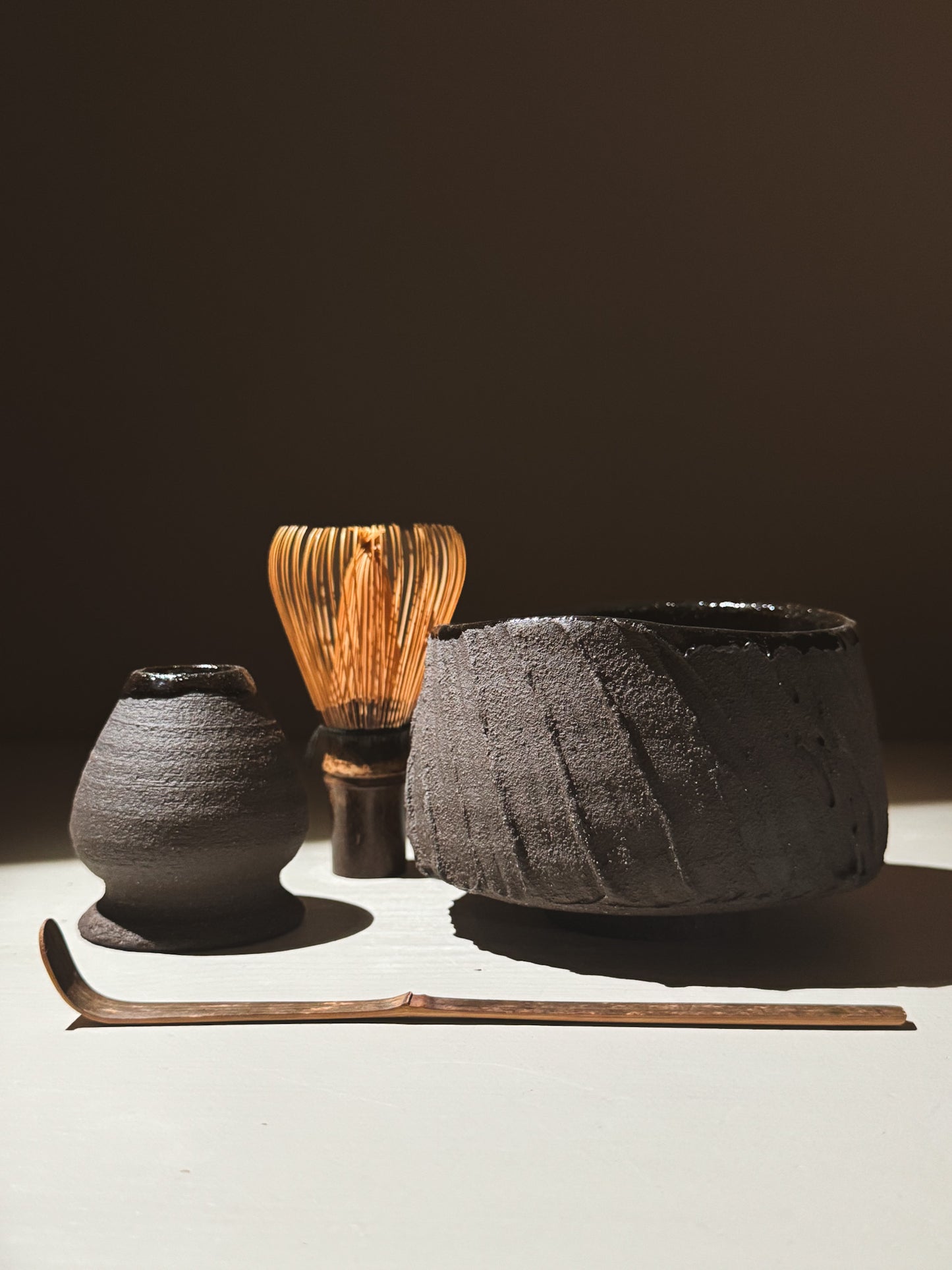 Kuro Matcha Bowl #07 with a matcha whisk, scoop, and teapot on a light surface, showcasing earthy textures.