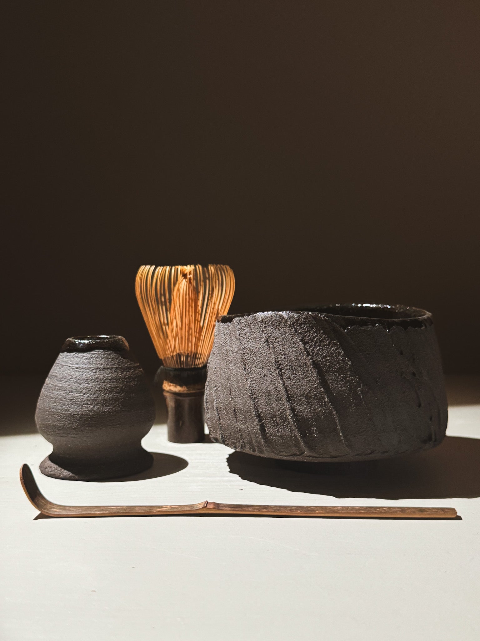 Kuro Matcha Bowl #07 with a matcha whisk, scoop, and teapot on a light surface, showcasing earthy textures.