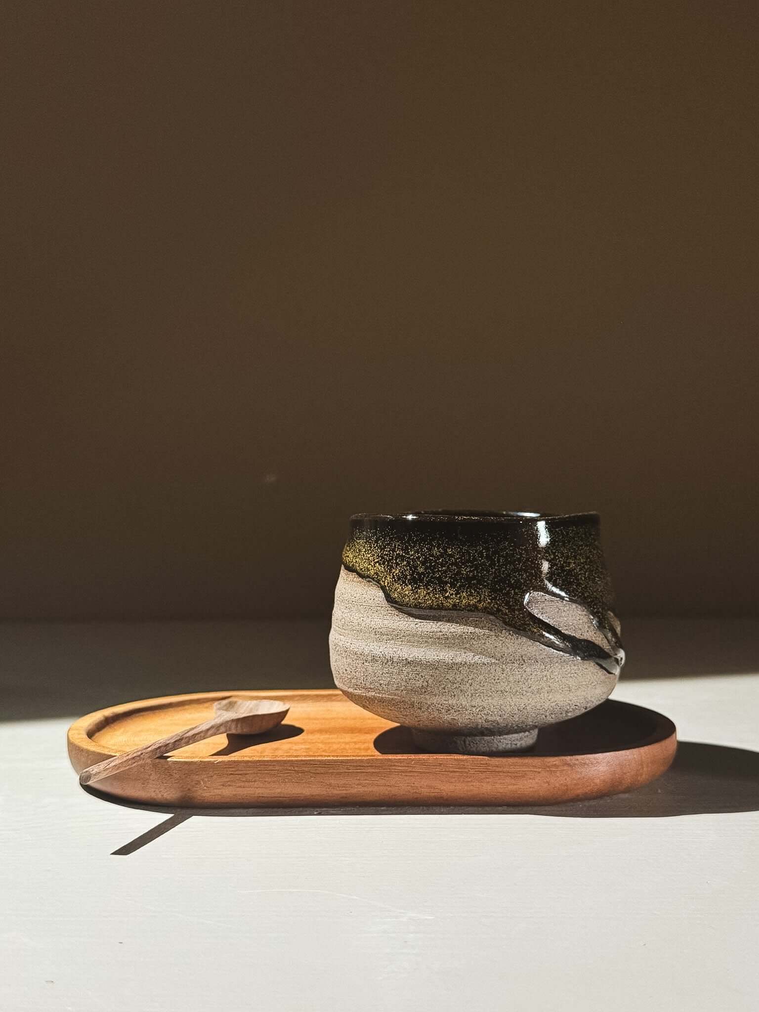 Yunomi Cup - Kuro Ash #01 on a wooden tray with a spoon, showcasing its glossy black glaze and natural finish.