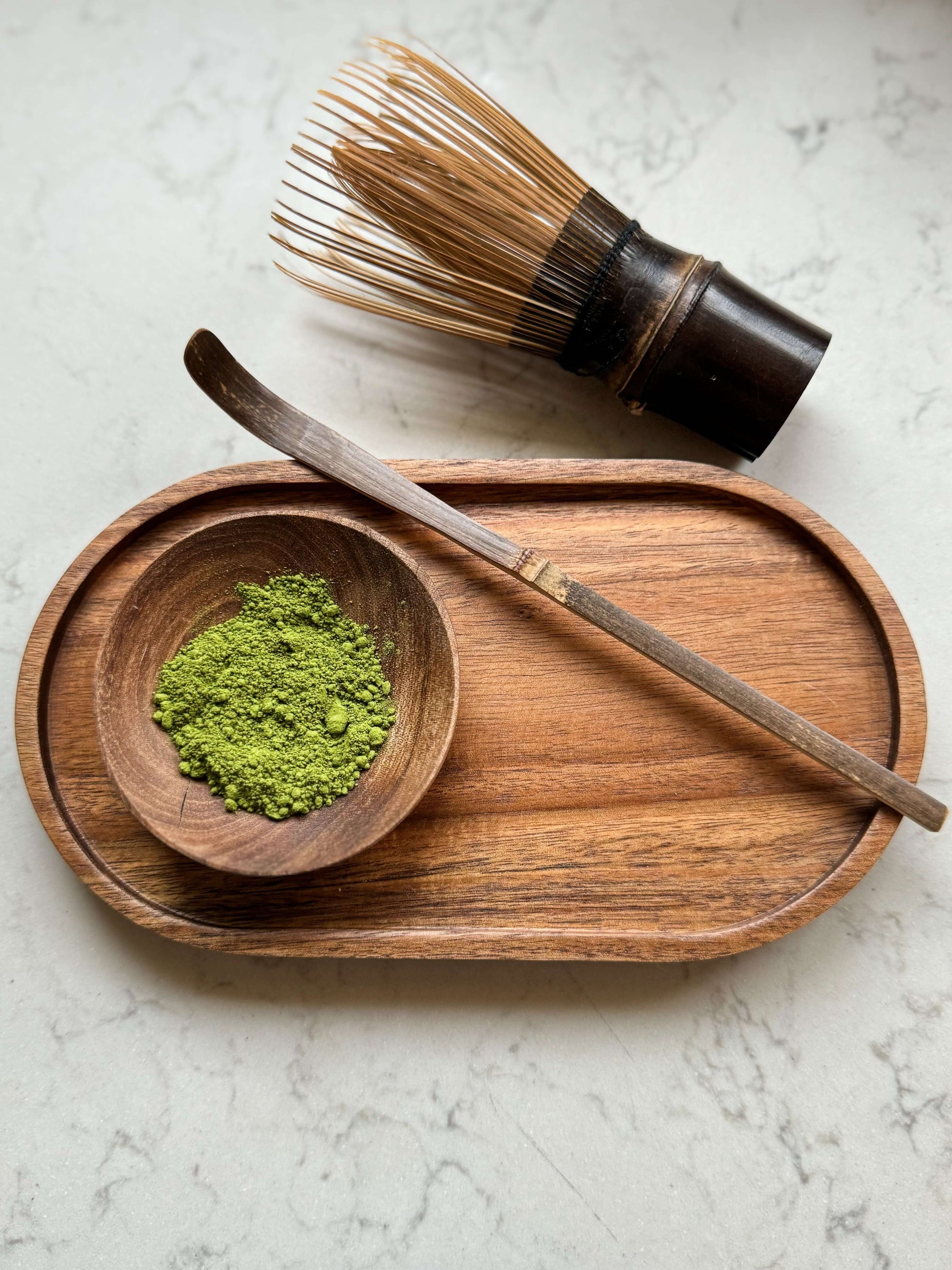 Dark bamboo matcha whisk (Chasen) on a wooden tray with matcha powder and spoon, showcasing natural elegance.