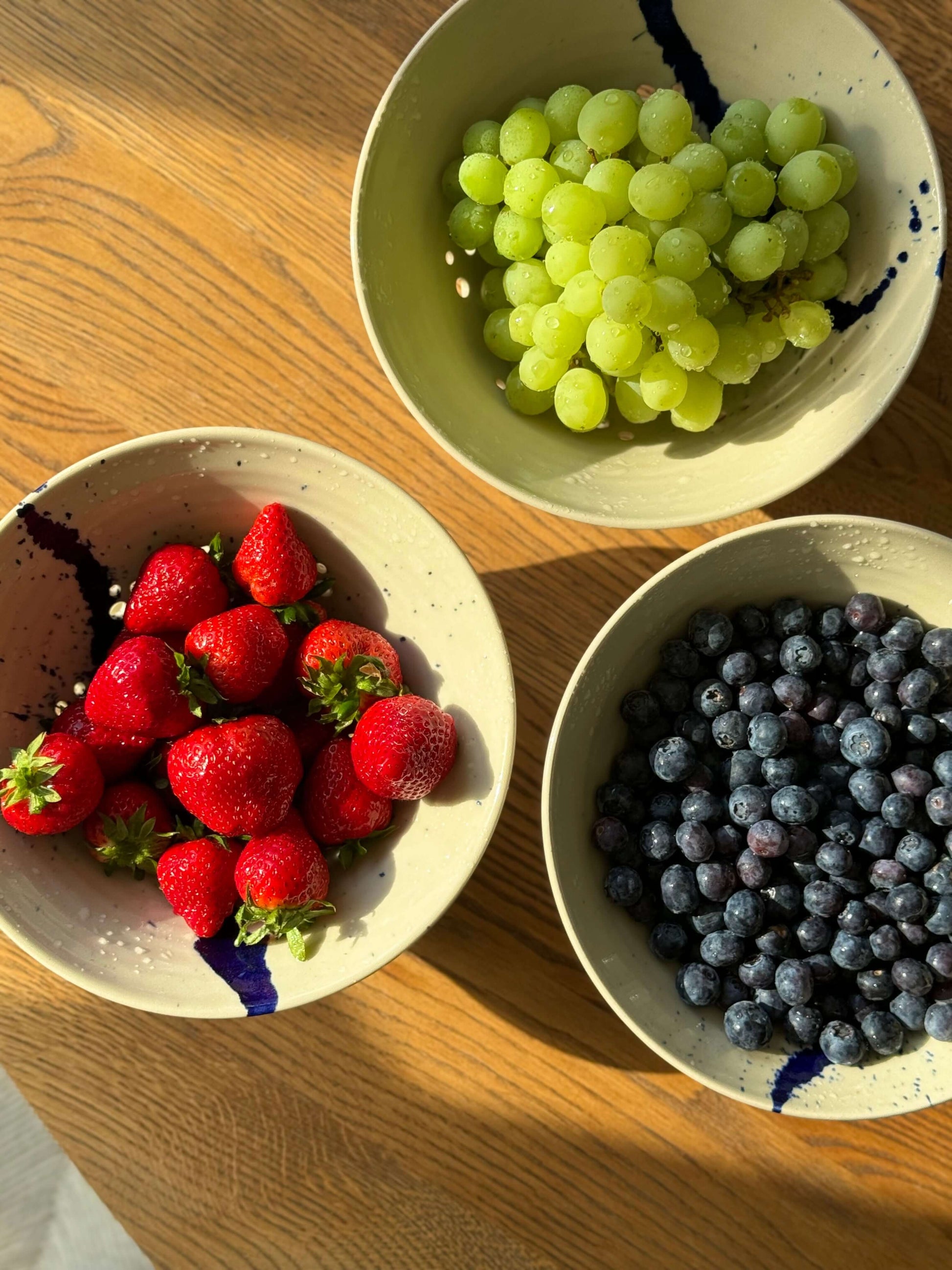 Strawberries, blueberries, and grapes in Gosu Series sculptural berry bowls with cobalt blue splashes, placed on a wooden table.