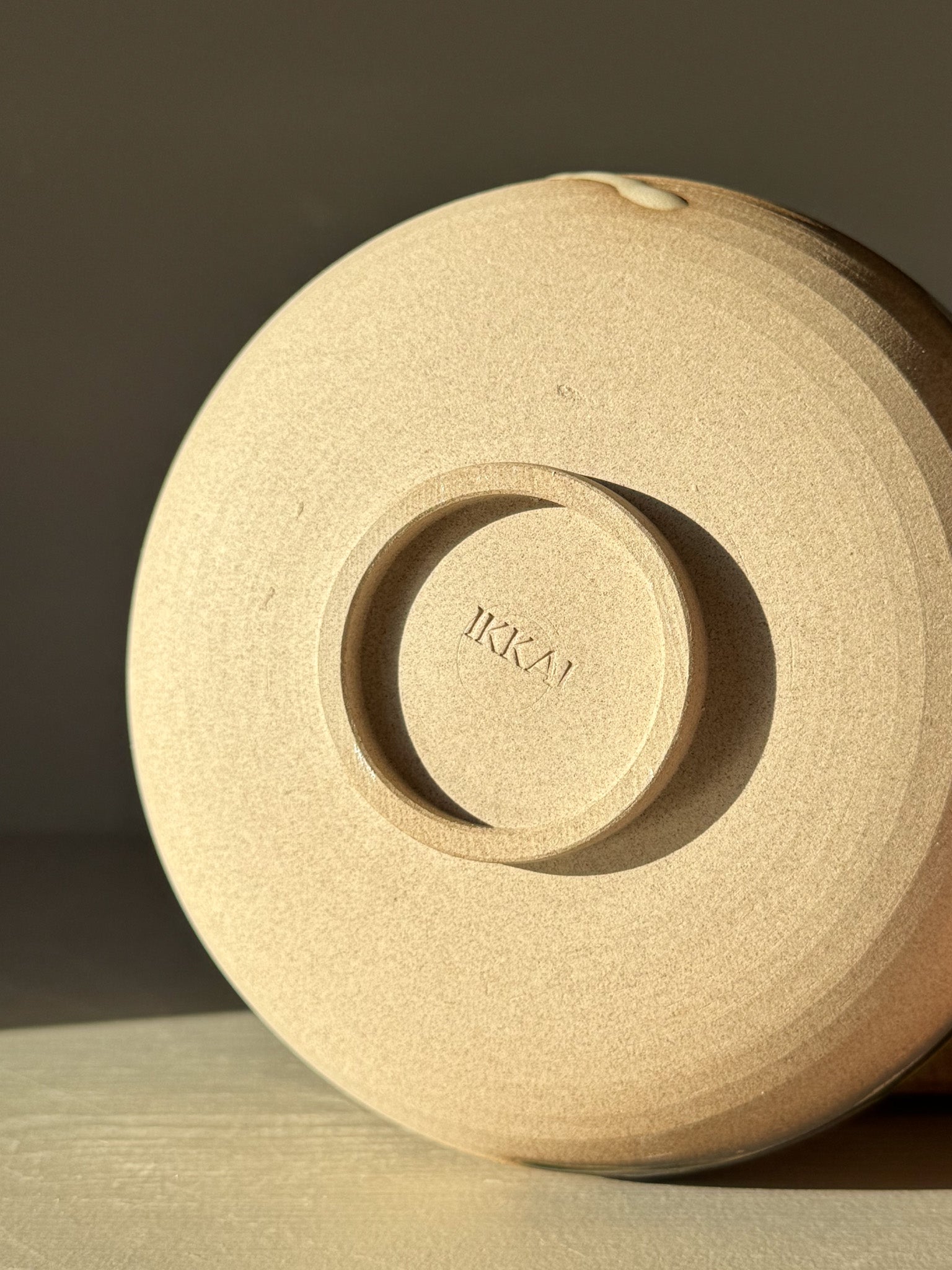 Bottom view of a ceramic bowl with "IKAI" stamped on the base, showcasing detailed craftsmanship and smooth finish in toffee-colored clay.