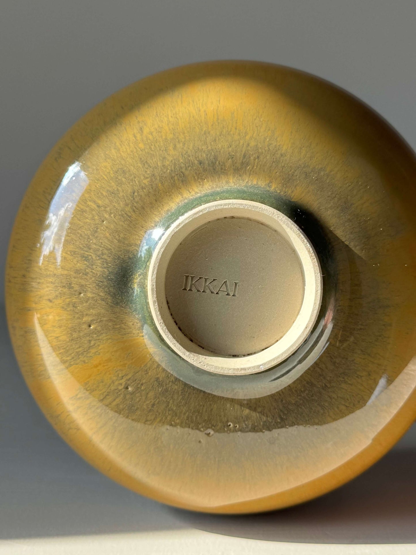 Base of a ceramic item with 'IKKAI' imprint, featuring a glossy gradient finish in yellow and brown shades.