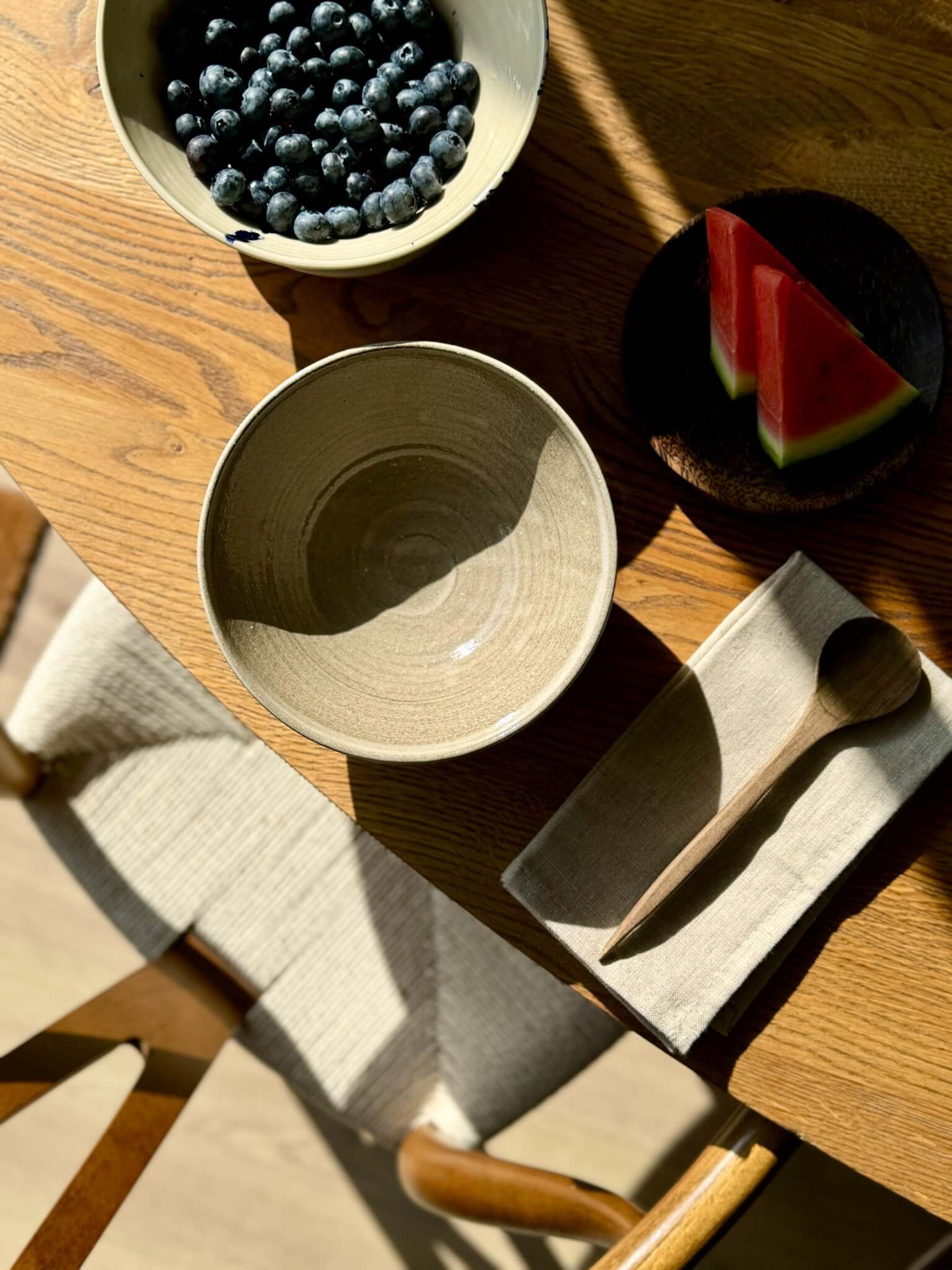 Bowl – Yuki Series Dark Clay handmade ceramic bowl on a wooden table next to fruit and spoon, showcasing its wide open shape and natural finish.