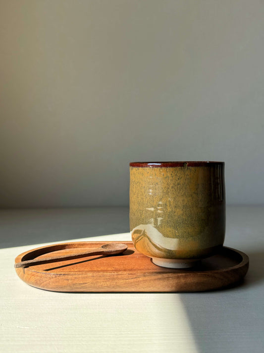 Handmade flower yunomi cup with tenmoku glaze, featuring amber, rust, and olive tones, on a wooden tray in natural light.