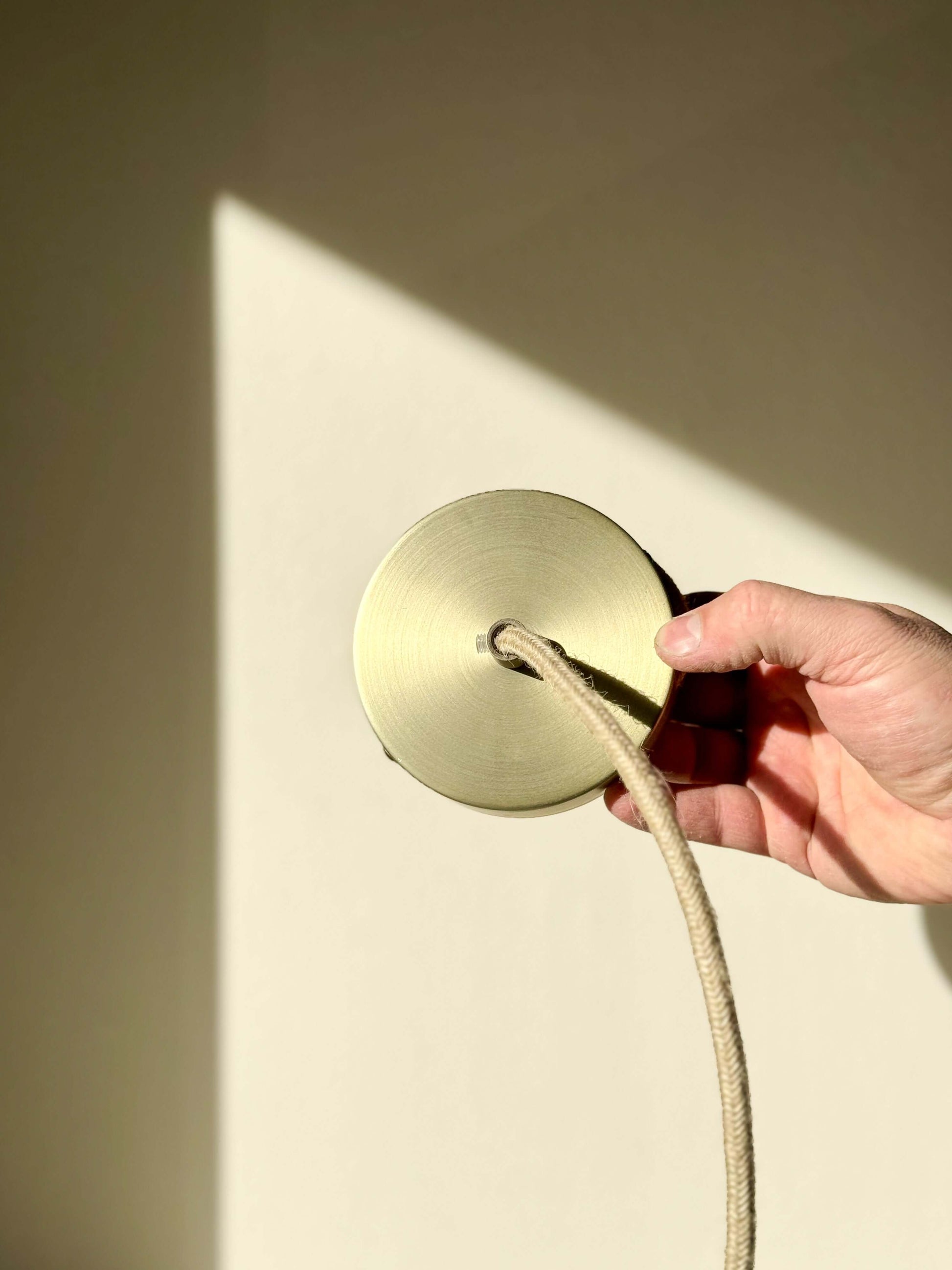 Close-up of a hand holding a round brass ceiling plate with a rope cord attached, illuminated by sunlight.