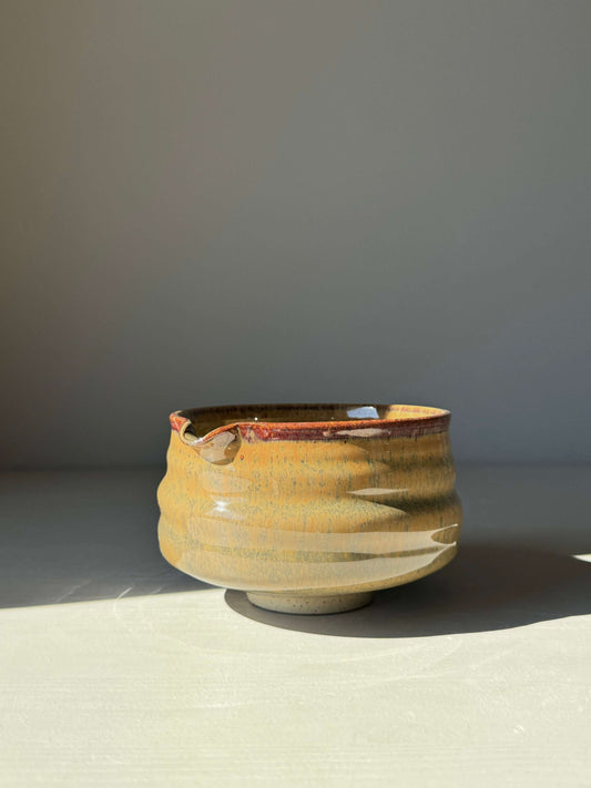 Hand-thrown matcha bowl with spout and tenmoku glaze, featuring carved grooves and a blend of brown and green hues under sunlight.