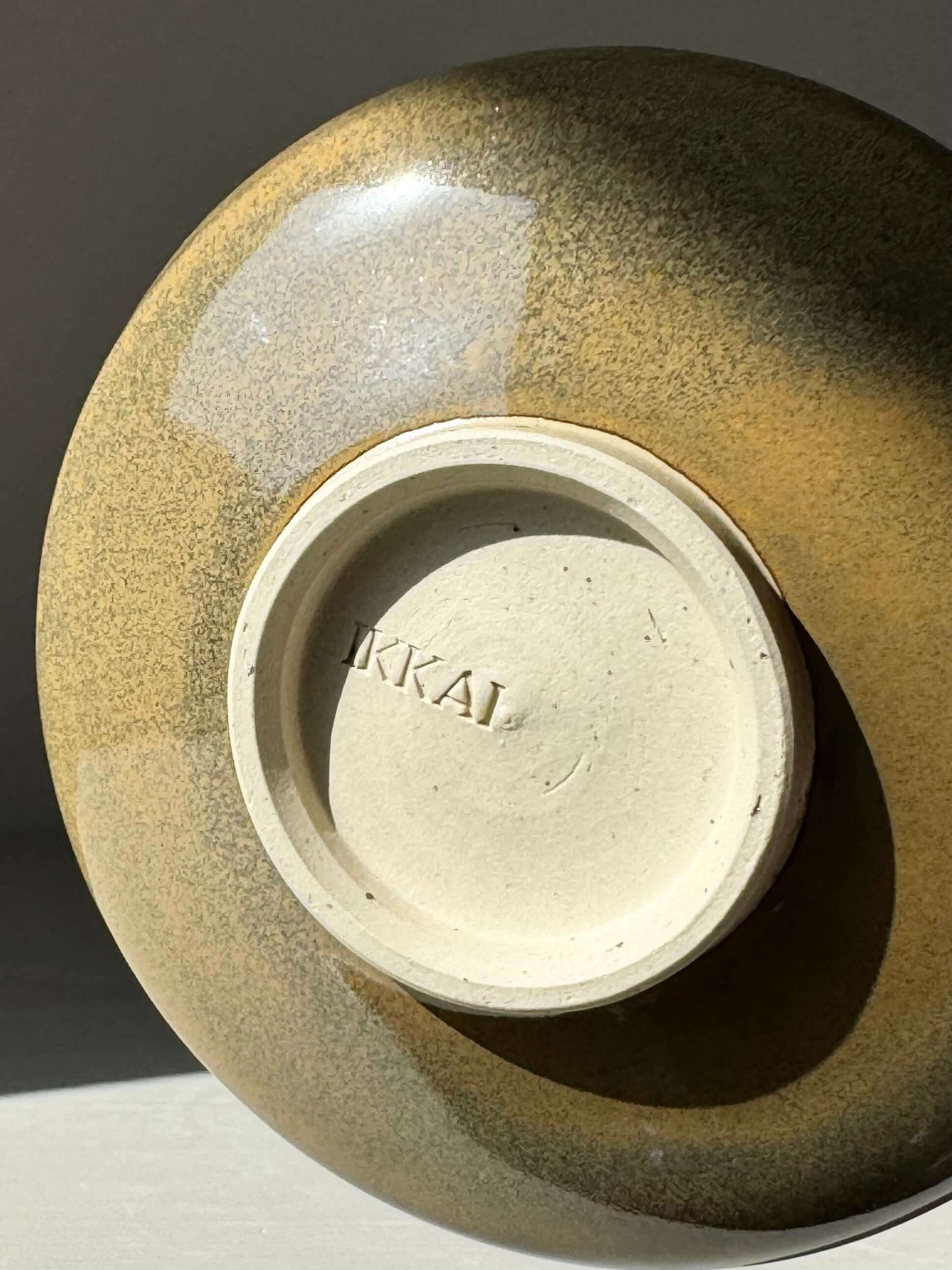Base of handmade Matcha bowl with tenmoku glaze, showing carved grooves and IKAI mark, in rich brown and soft green hues.