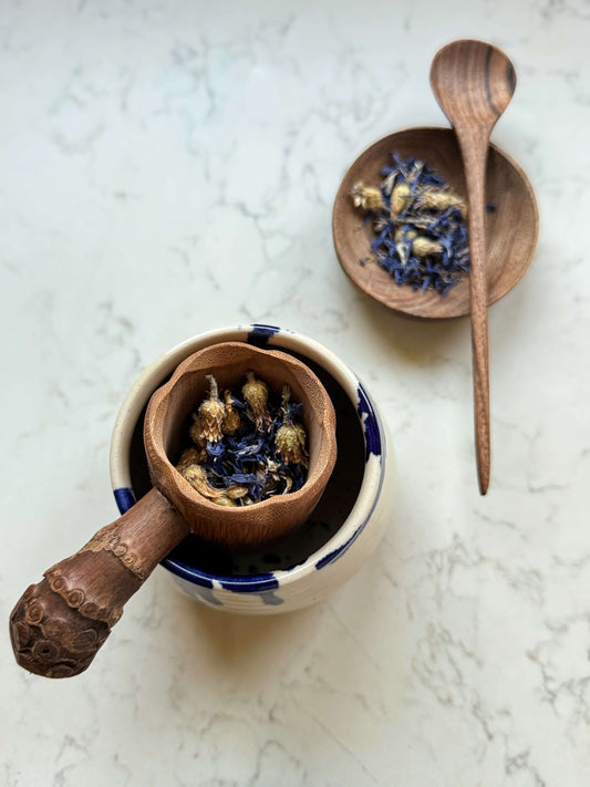 Handmade bamboo tea strainer with root handle placed on a mug, featuring dried flowers, next to a wooden spoon and bowl.