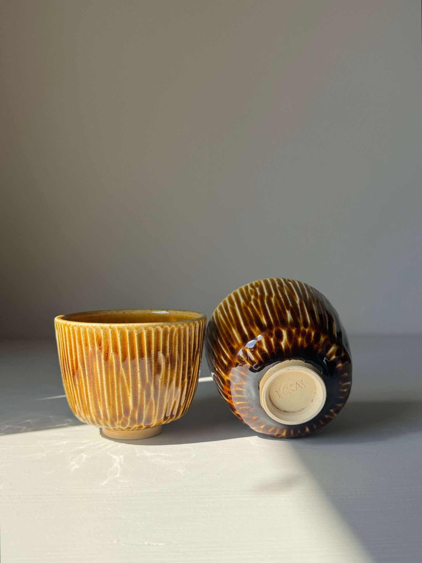 Handmade ceramic Carved Cups - Dark Honey with deep honey glaze, featuring grooved design and smooth rim, resting in sunlight.