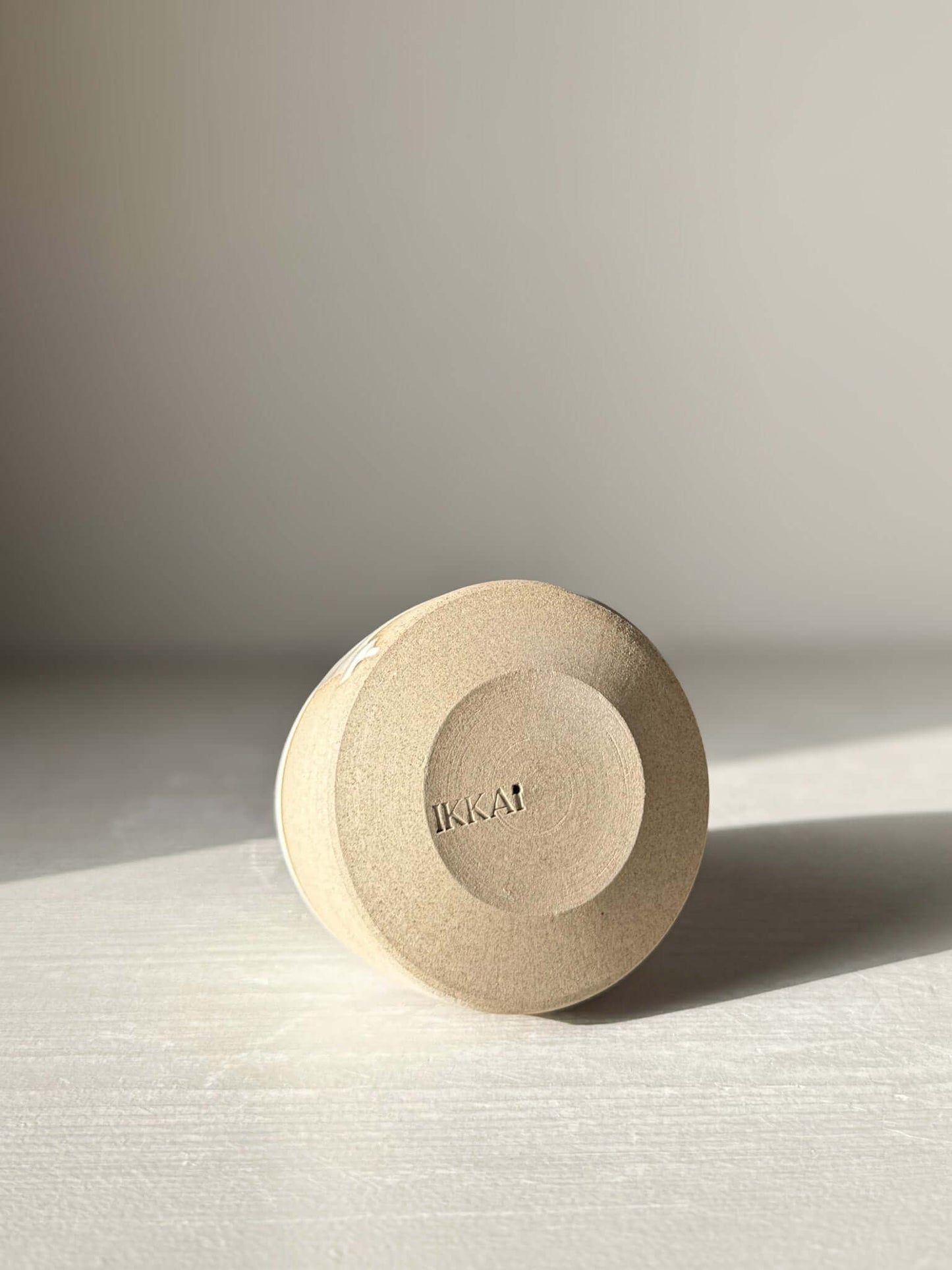 Bottom view of a handmade ceramic object with "IKKAI" imprint, showcasing a minimal design. Soft lighting highlights the light texture.