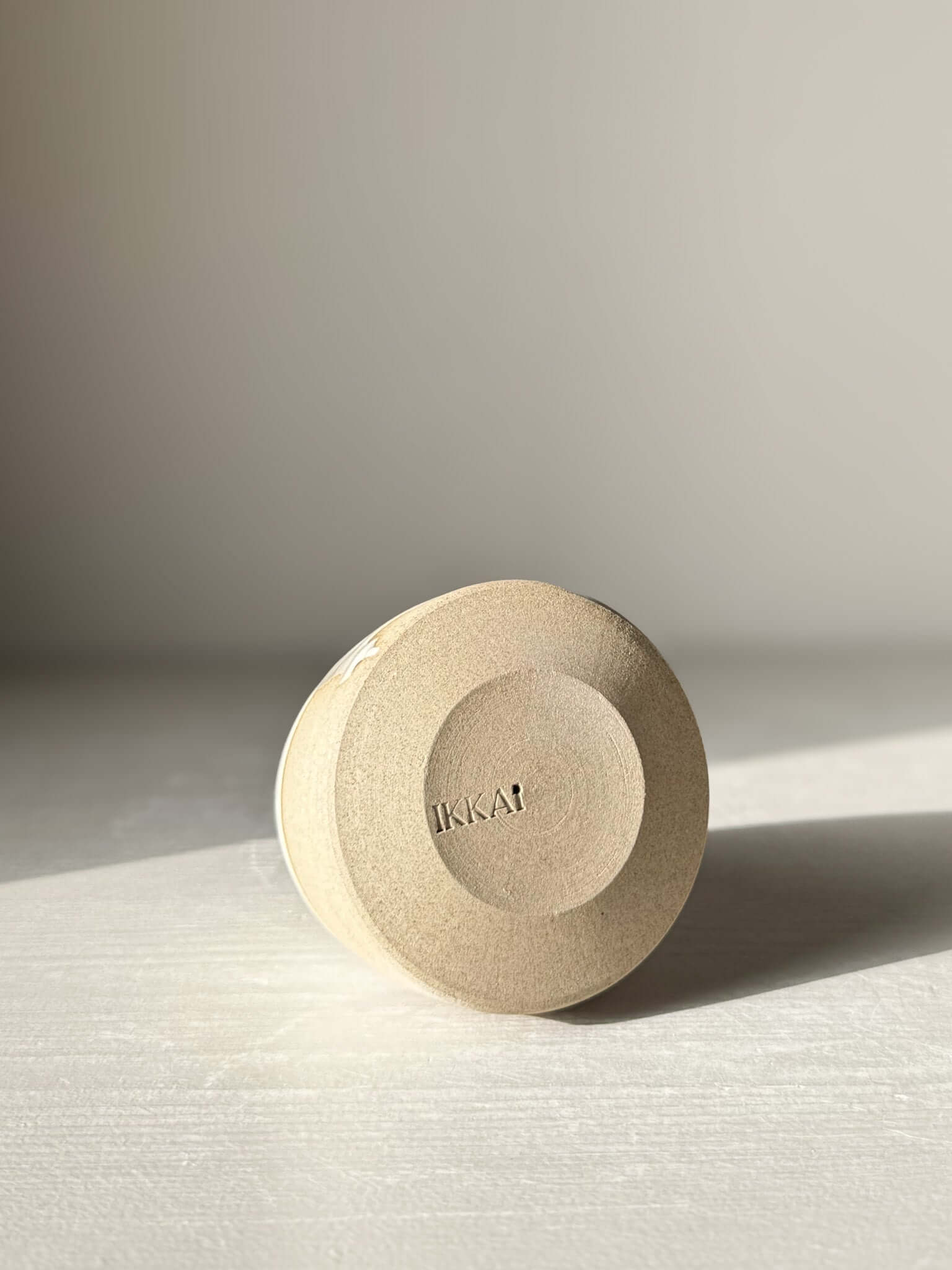 Bottom view of a handmade ceramic object with "IKKAI" imprint, showcasing a minimal design. Soft lighting highlights the light texture.