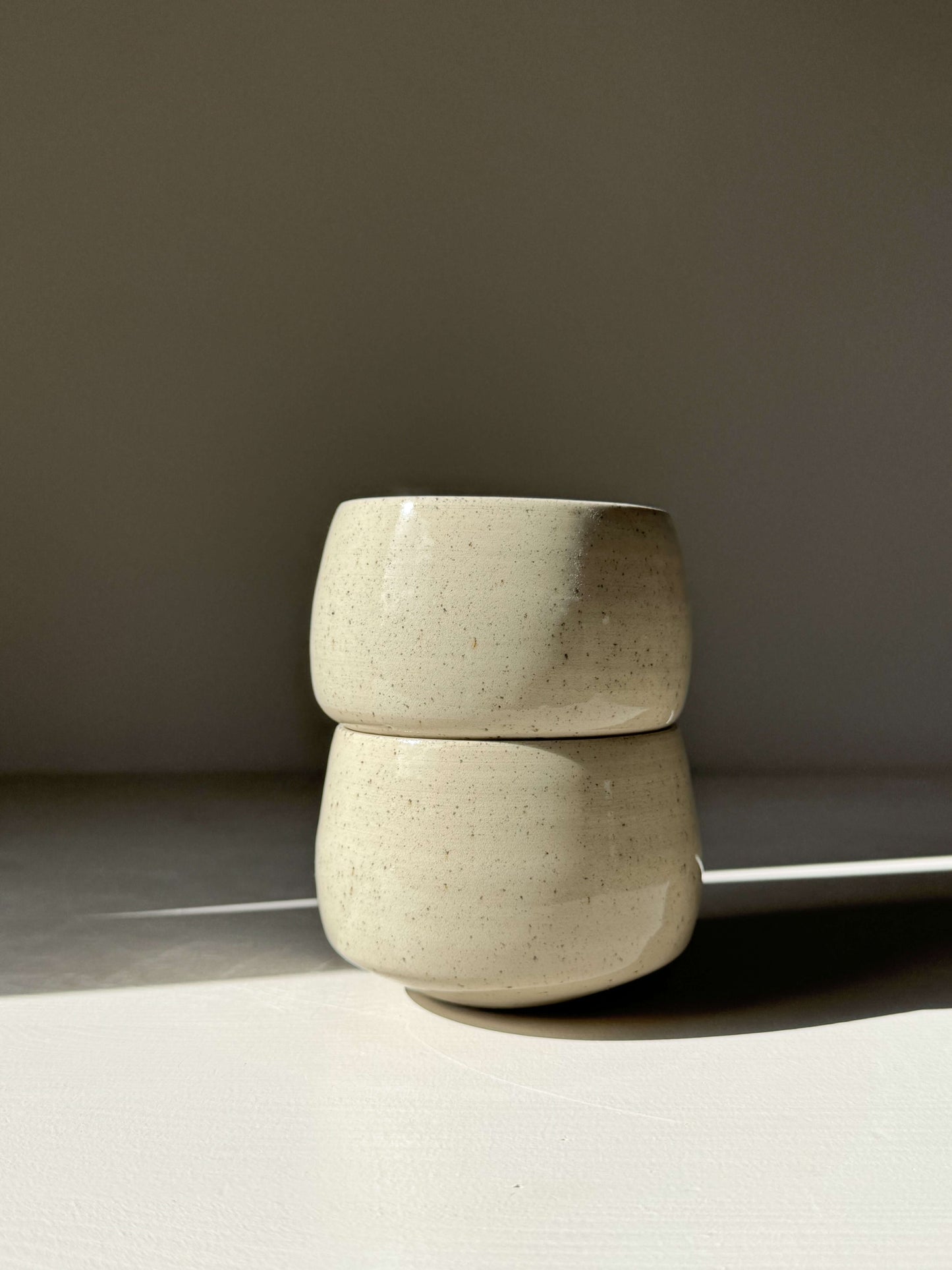 Stacked handmade ceramic cups with speckled clear glaze under soft light. Product: Cups - Speckled Clear Glaze, soft rounded shape.