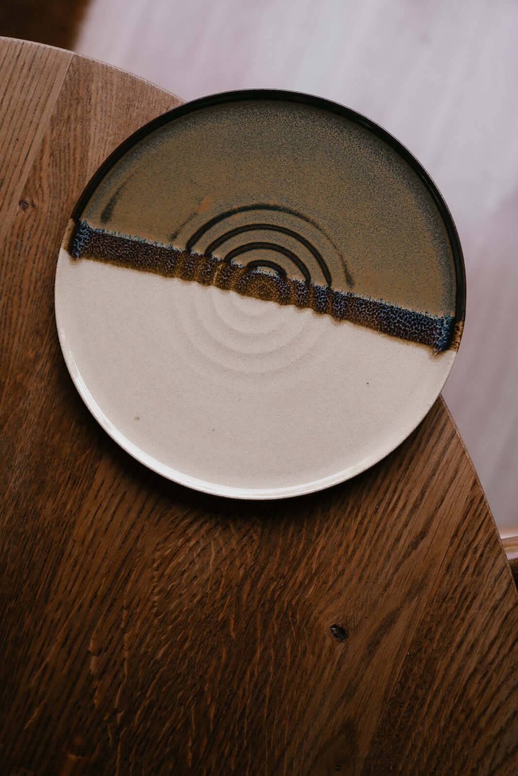 Handmade ceramic Flat Round Breakfast Plate – Inyo with half glossy tenmoku and toffee-colored stoneware on a wooden table.