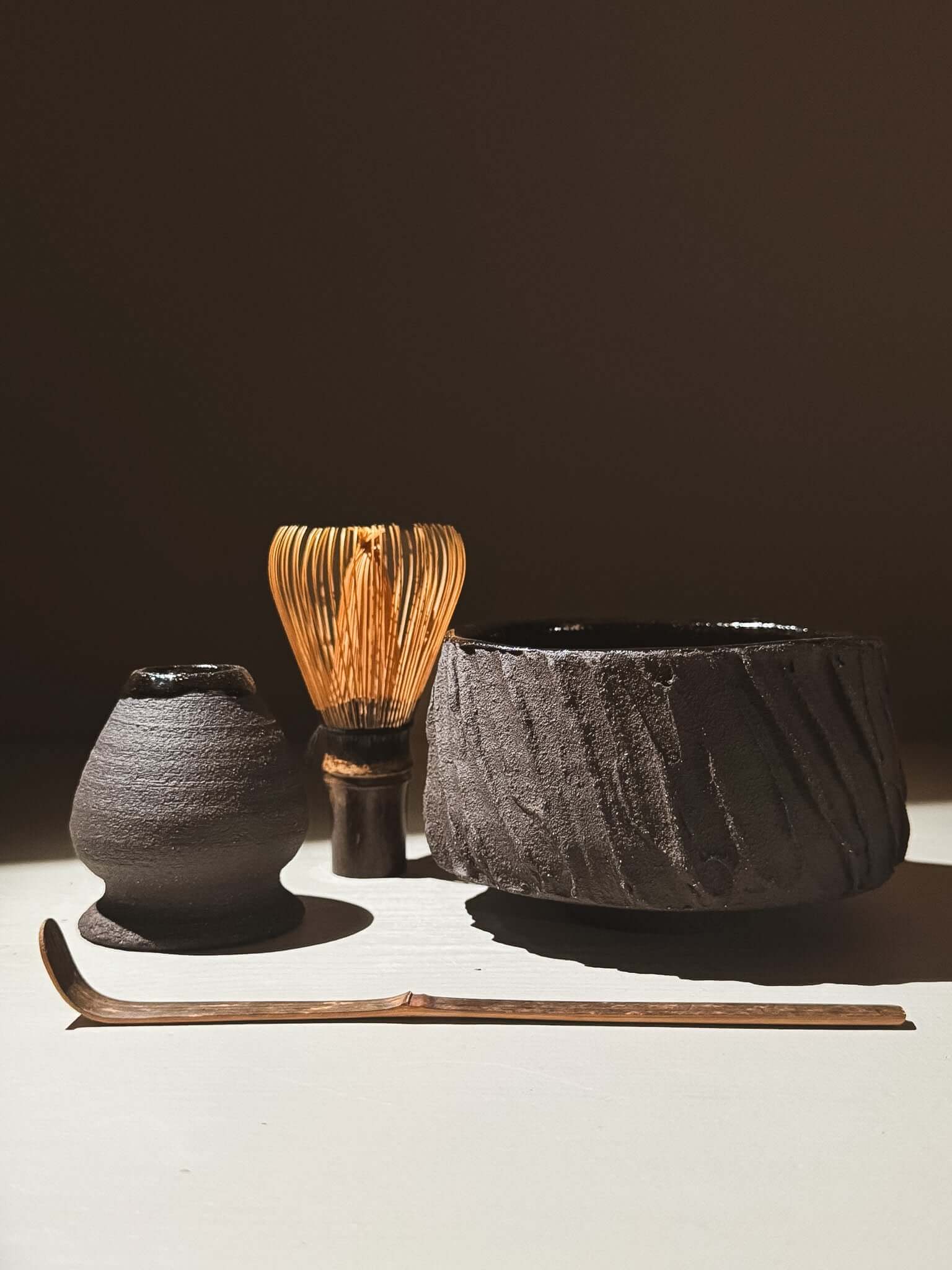 Kuro Matcha Bowl #06 set with whisk, bowl, and scoop on a light surface, showcasing earthy textures and glossy interior.